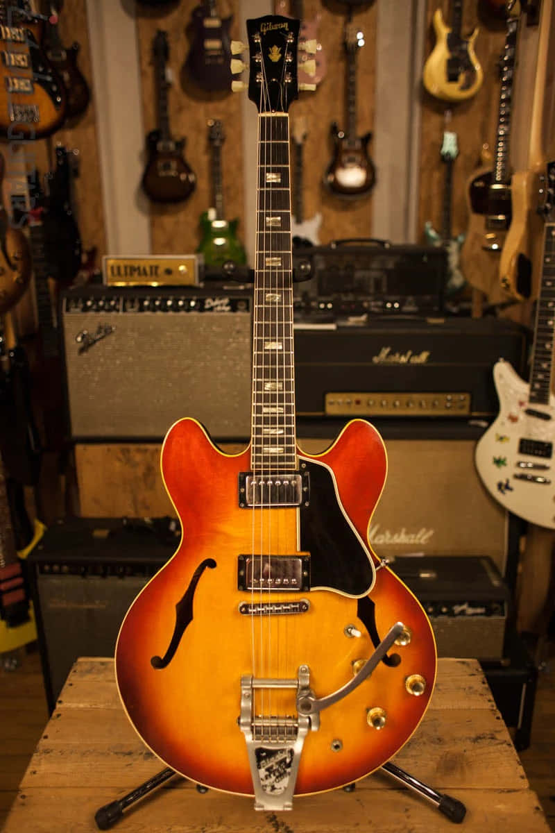 Guitar Aesthetic 桌布 壁紙 — 一把漸變色Gibson ES-335電吉他放在木製支架上，位於音樂店中央，周圍有擴音器和掛著的吉他。