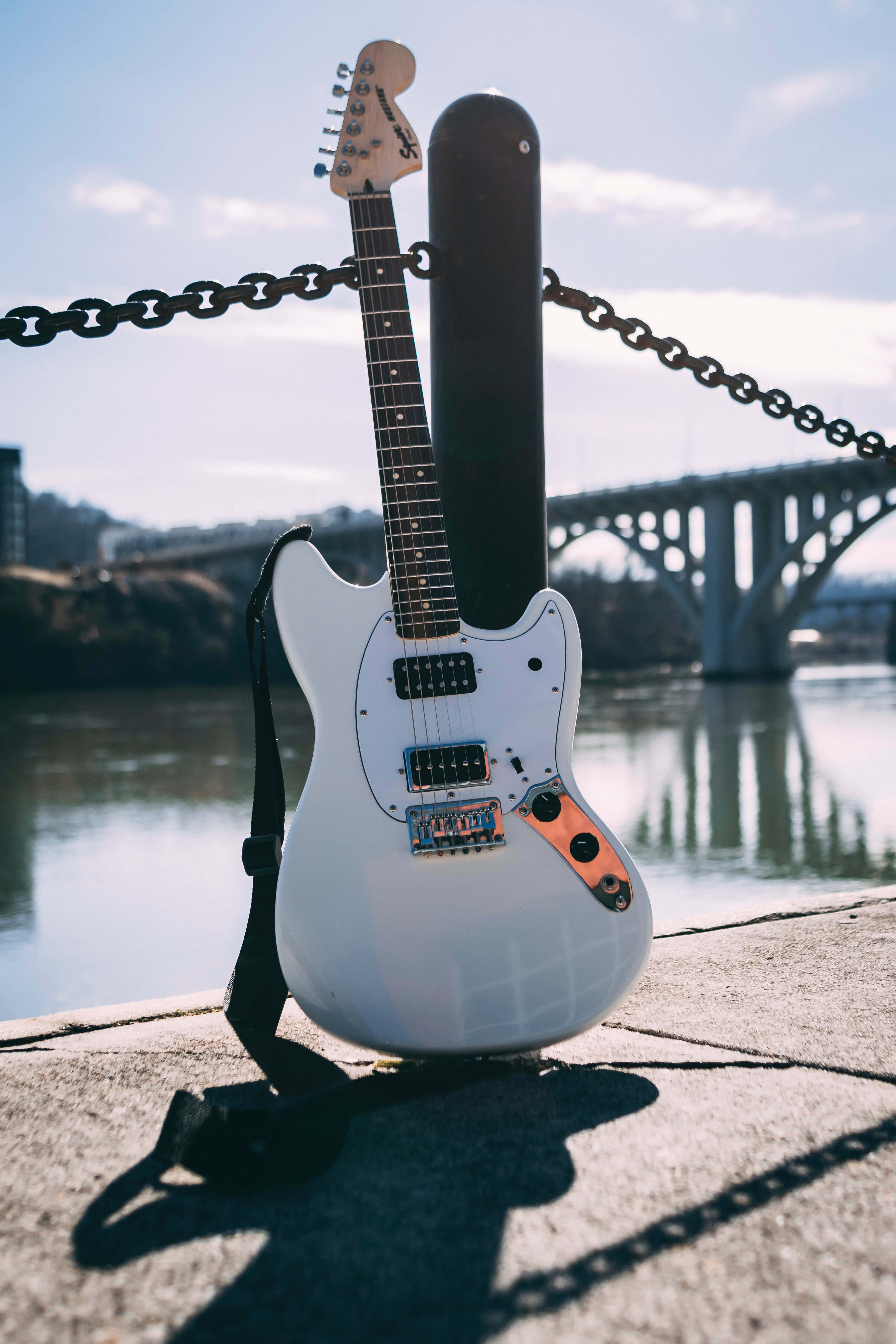 Guitar Aesthetic 桌布 壁紙 — 一把白色電吉他連黑色琴盒靠在河邊的鐵絲網圍欄上，背景有座橋。