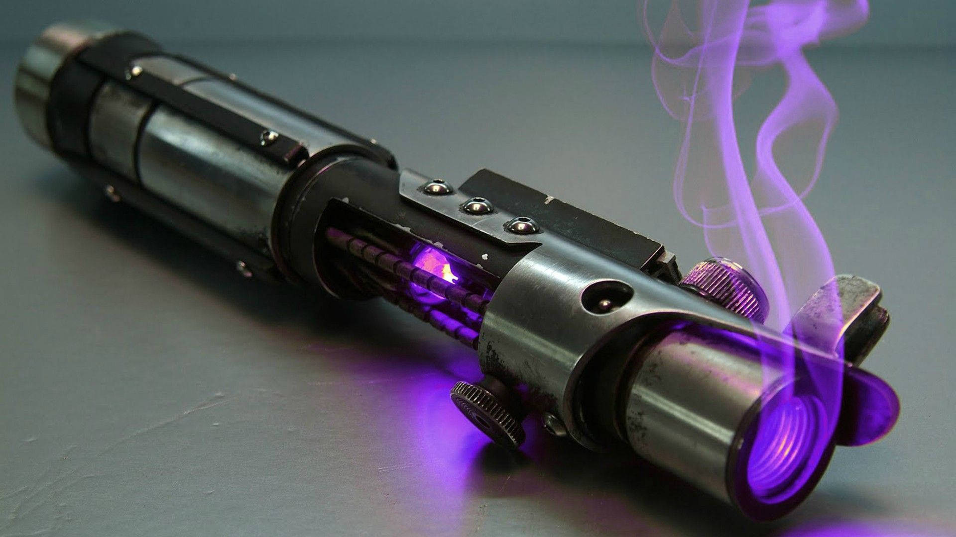 Lightsaber 桌布 壁紙 — 一把紫色光劍，帶有發光的劍刃和從劍柄升起的煙霧，放置在灰色表面上。