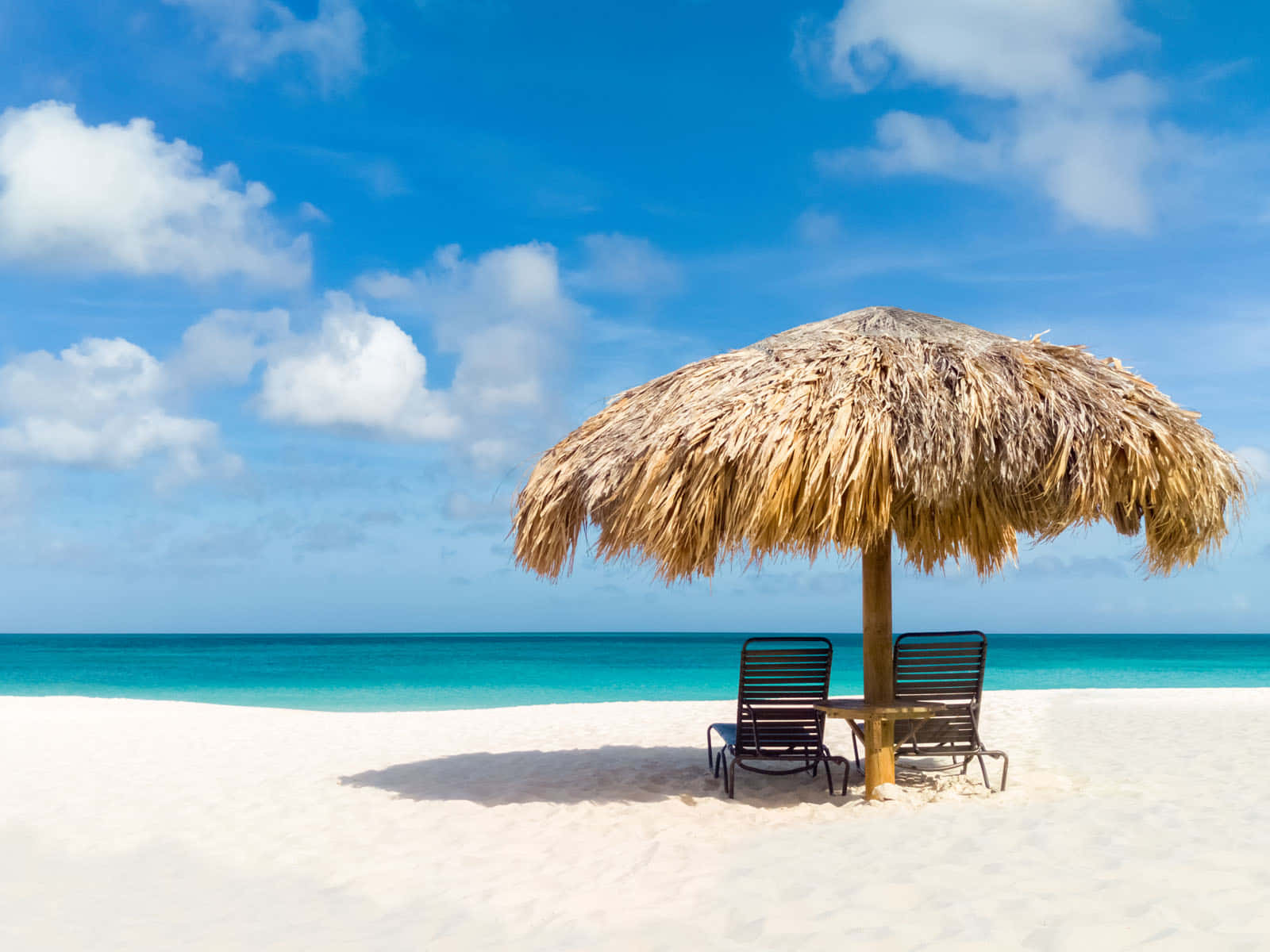 Aruba 桌布 壁紙 — 一把茅草遮陽傘為純白沙灘上的兩張躺椅遮蔭，背景是晴朗藍天和 fluffy 白雲。