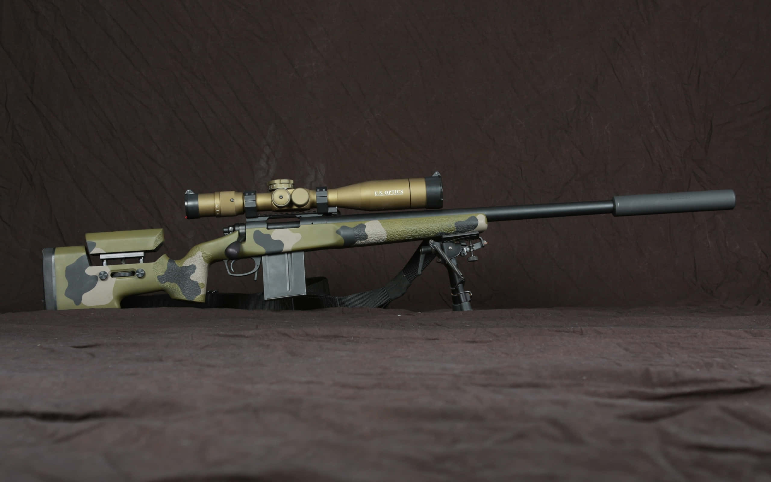 Rifle 桌布 壁紙 — 一把迷彩狙擊步槍配金色瞄準鏡，架在兩腳架上，背景為深棕色布料。