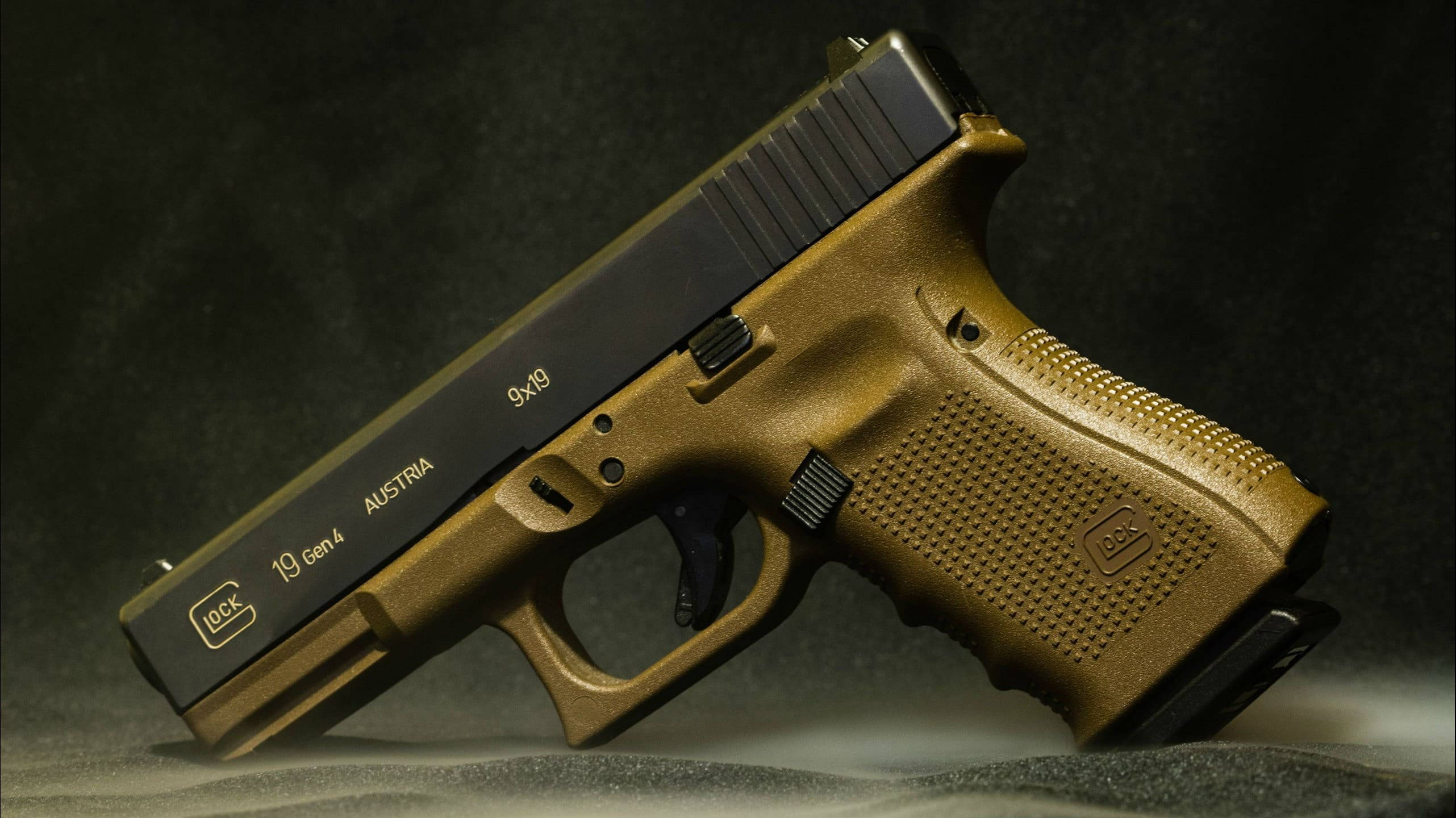 克拉克 桌布 壁紙 — 一把金色Glock 19 Gen4手槍對角放置在深色紋理表面上，在戲劇性的工作室燈光下。