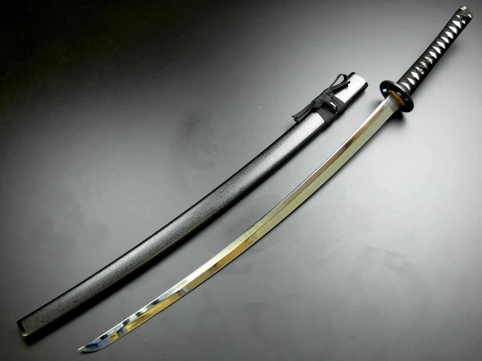 Samurai Sword 桌布 壁紙 — 一把閃亮的武士刀，帶有拋光刀片和黑色紋理刀鞘，斜放在深灰色表面上。