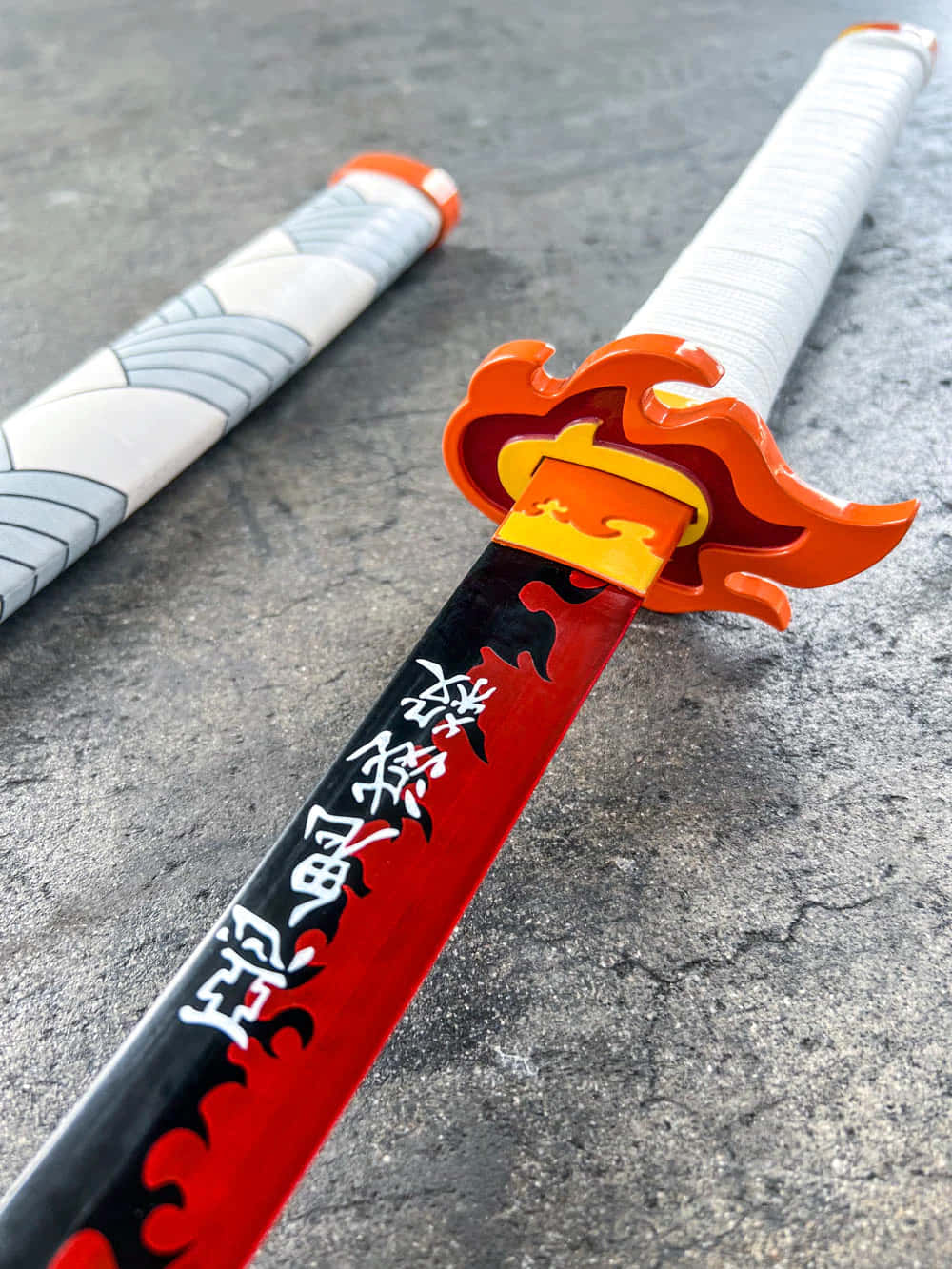 Rengoku Sword 桌布 壁紙 — 一把鮮紅與黑色的劍，帶有橙色火焰劍柄，擱在其帶有圖案的劍鞘旁，置於有紋理的灰色表面上。