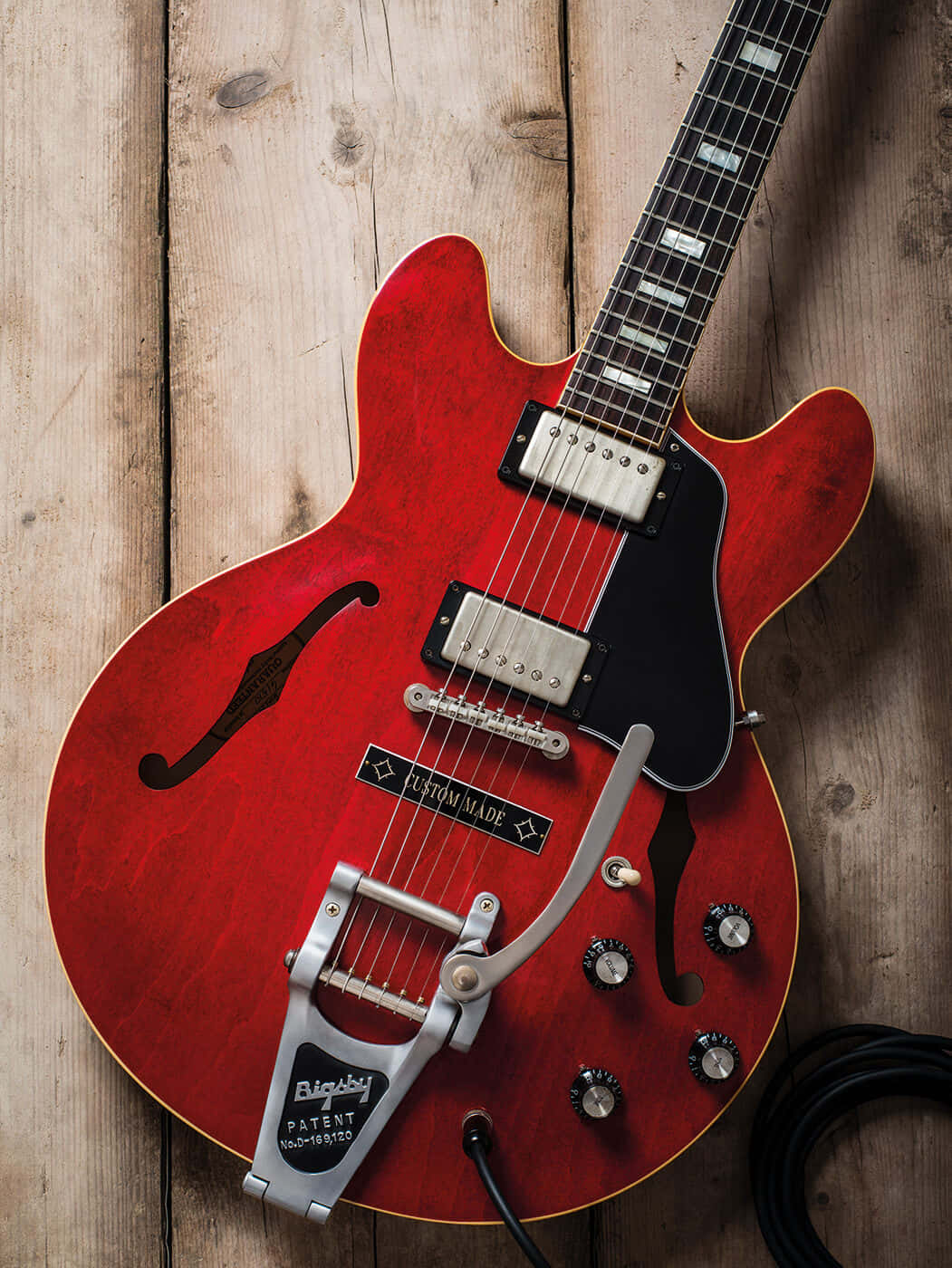 Guitar Aesthetic 桌布 壁紙 — 一把鮮紅色的電吉他，配有 Bigsby 顫音搖把，擱在風化的木板上，其光亮的表面反射出溫暖的光線。