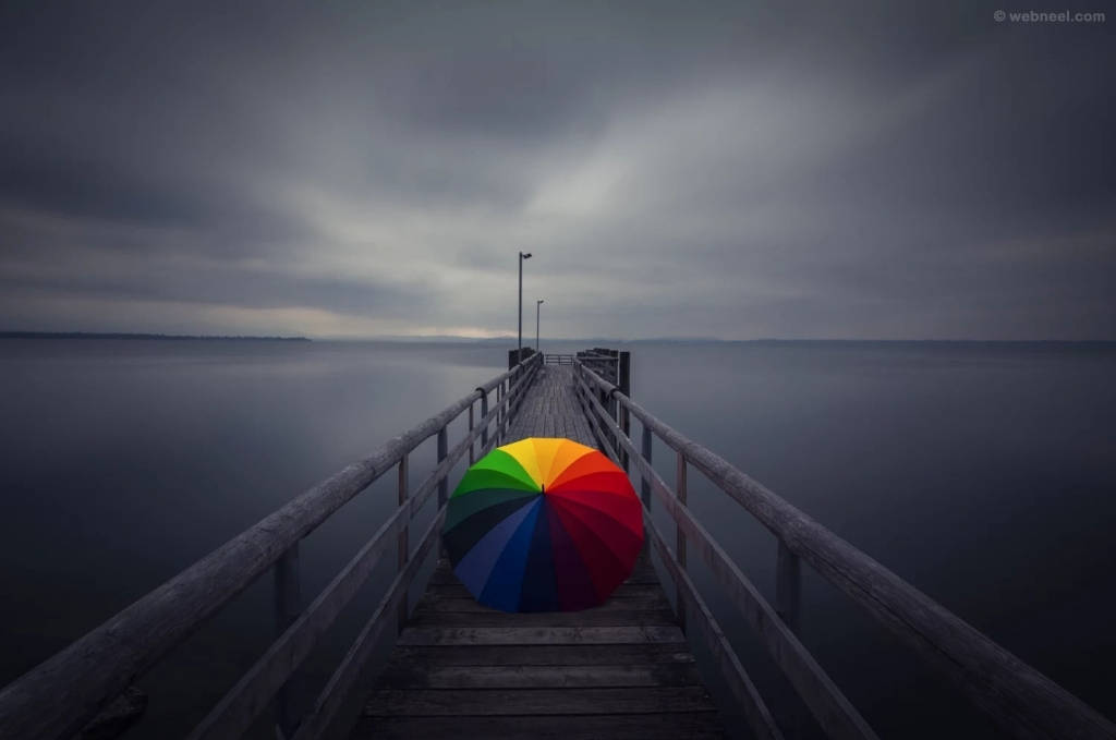 Umbrella 桌布 壁紙 — 一把鮮豔的彩虹傘擱在一個風化的木碼頭上，碼頭延伸至陰天下一片霧濛濛的灰色湖泊。