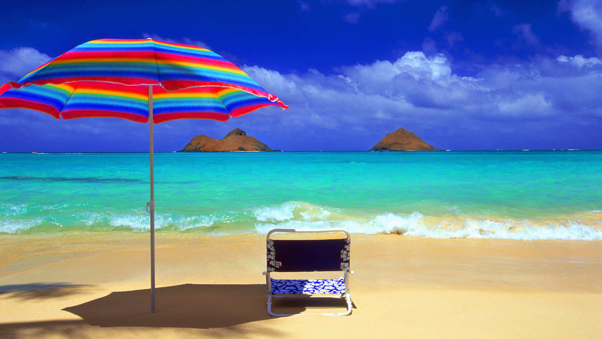 Beach Sunbed 桌布 壁紙 — 一把鮮豔的彩虹傘為金色沙灘上的圖案沙灘椅遮蔭，旁邊是碧綠的海浪，深藍的天空下遠處有火山島嶼。
