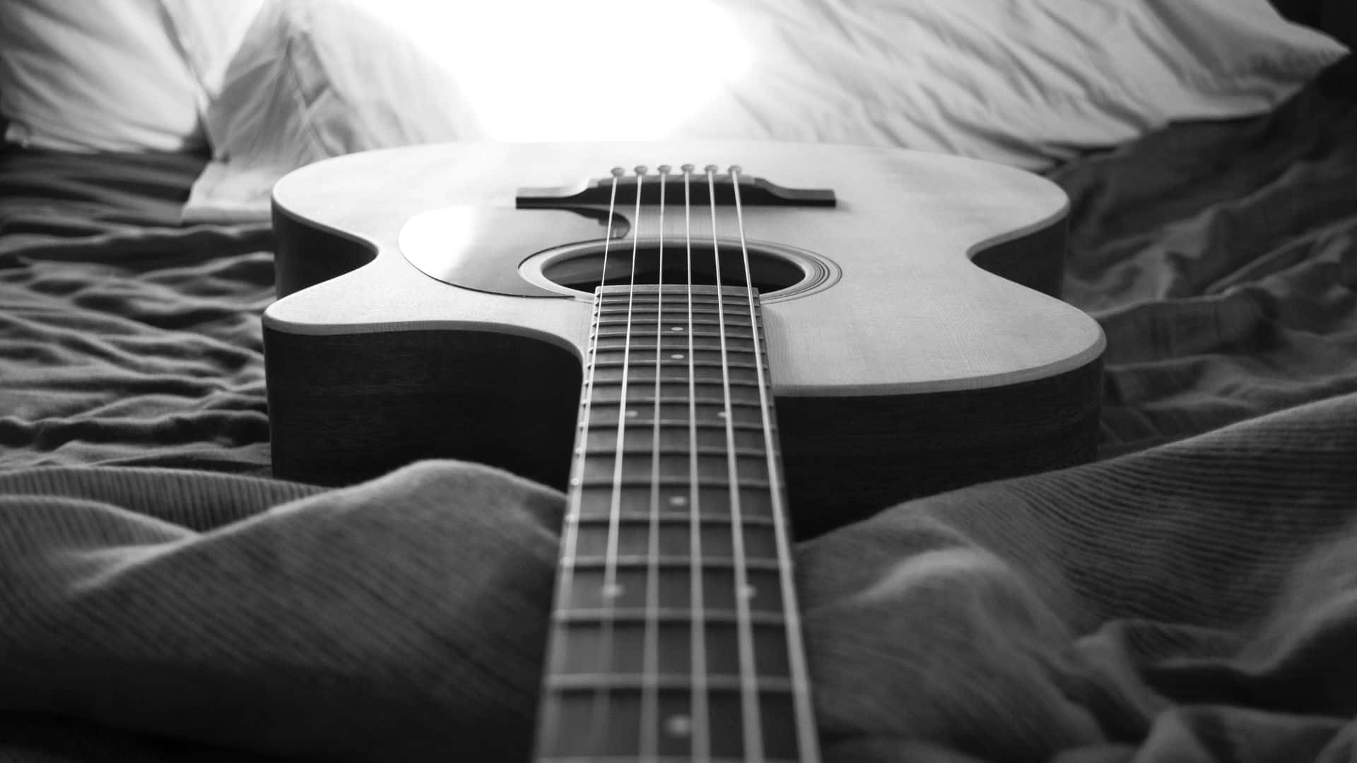 Black And White Guitar 桌布 壁紙 — 一把黑白原聲吉他放在皺巴巴的床單上，枕頭後透出柔和光線。