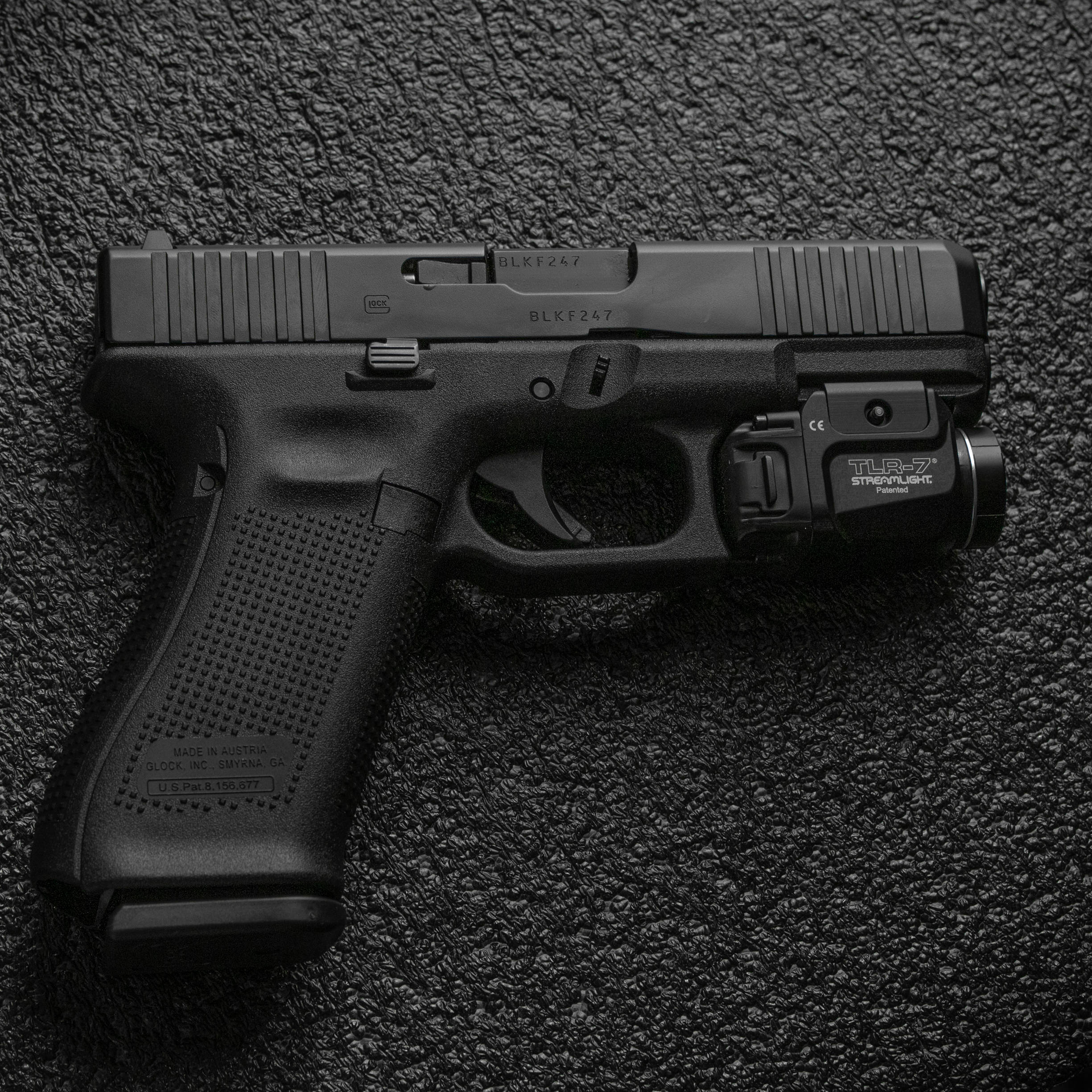 槍枝 桌布 壁紙 — 一把黑色 Glock 手槍配備 TLR-7 雷射瞄準器，放置在深色紋理表面上，呈現出鮮明、高對比度的攝影棚拍攝效果。
