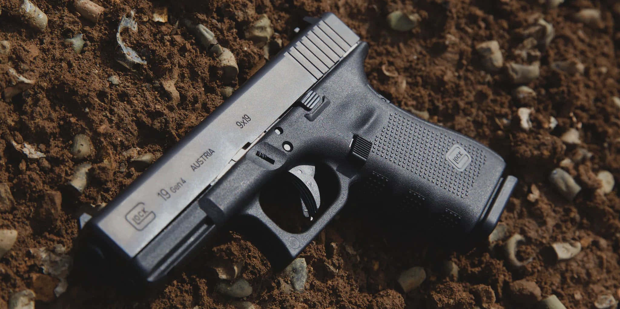 Glock 19 桌布 壁紙 — 一把黑色Glock 19 Gen4手槍在明亮日光下放置於粗糙的棕色泥土上，散落著小石子。