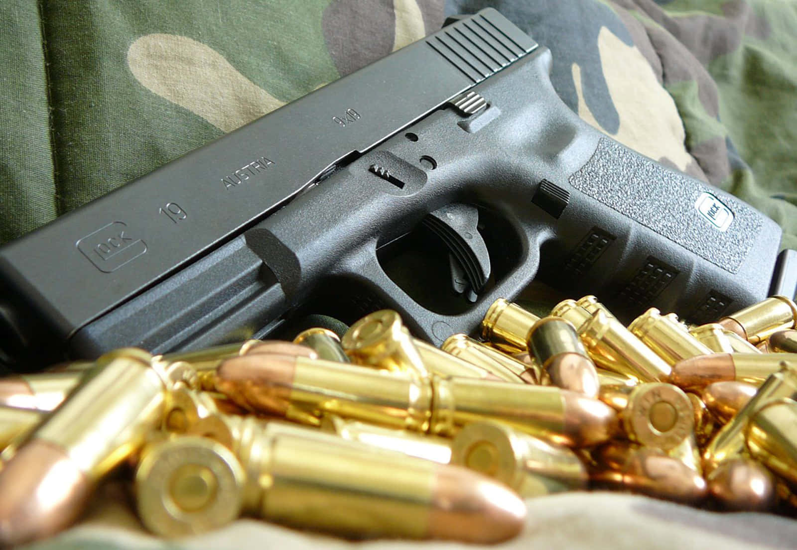 Glock 19 桌布 壁紙 — 一把黑色Glock 19手槍放在迷彩布料上，周圍散落著黃銅彈藥。