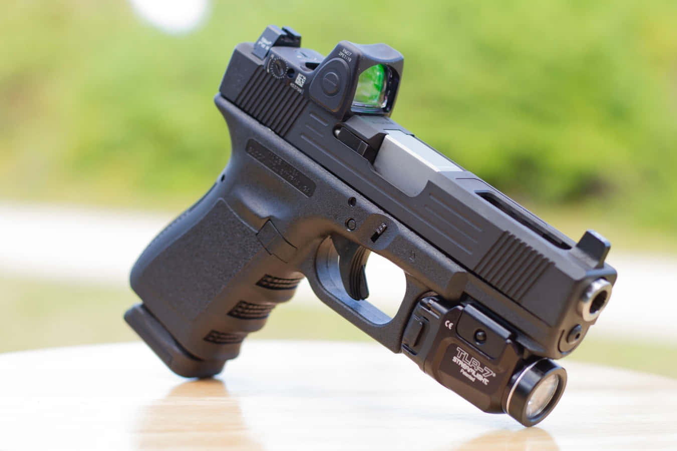 Glock 19 桌布 壁紙 — 一把黑色半自動手槍配備紅點瞄準器和戰術燈，放置在木質表面上，背景為模糊的綠色。