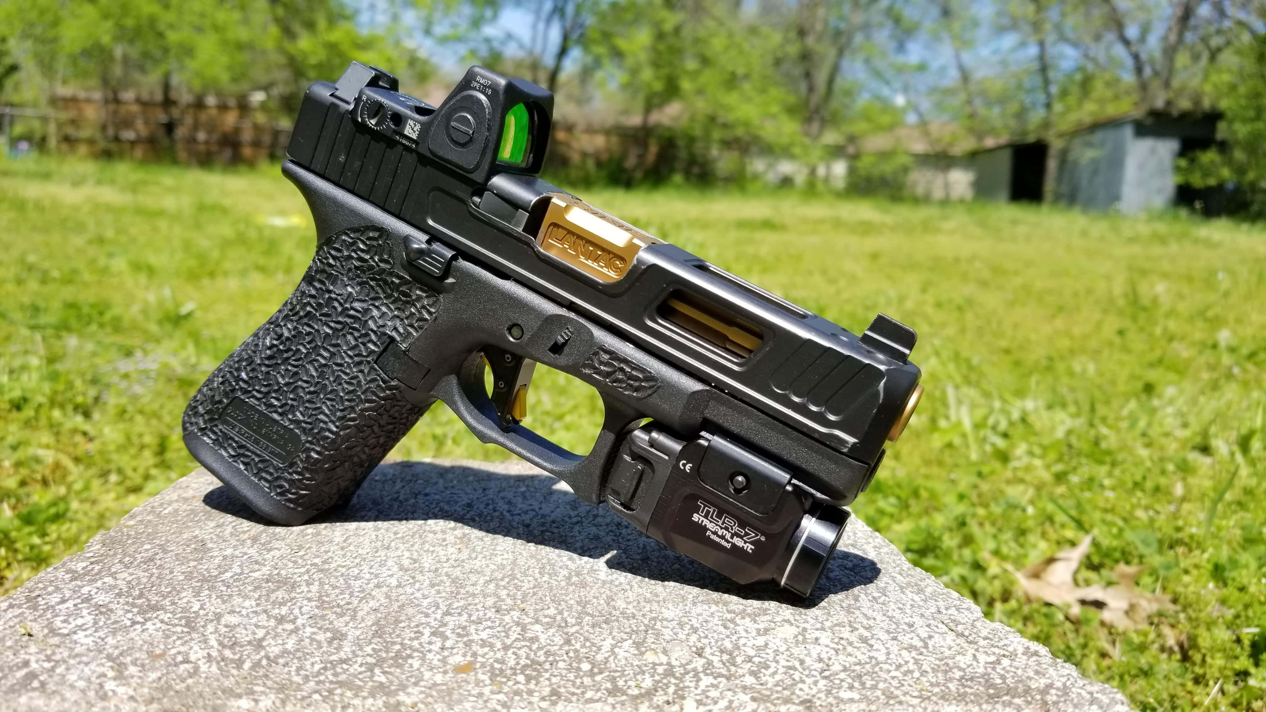 Glock 19 桌布 壁紙 — 一把黑色和金色的手槍配備紅點瞄準器，擱在草地庭院中一塊陽光照耀的石頭上。