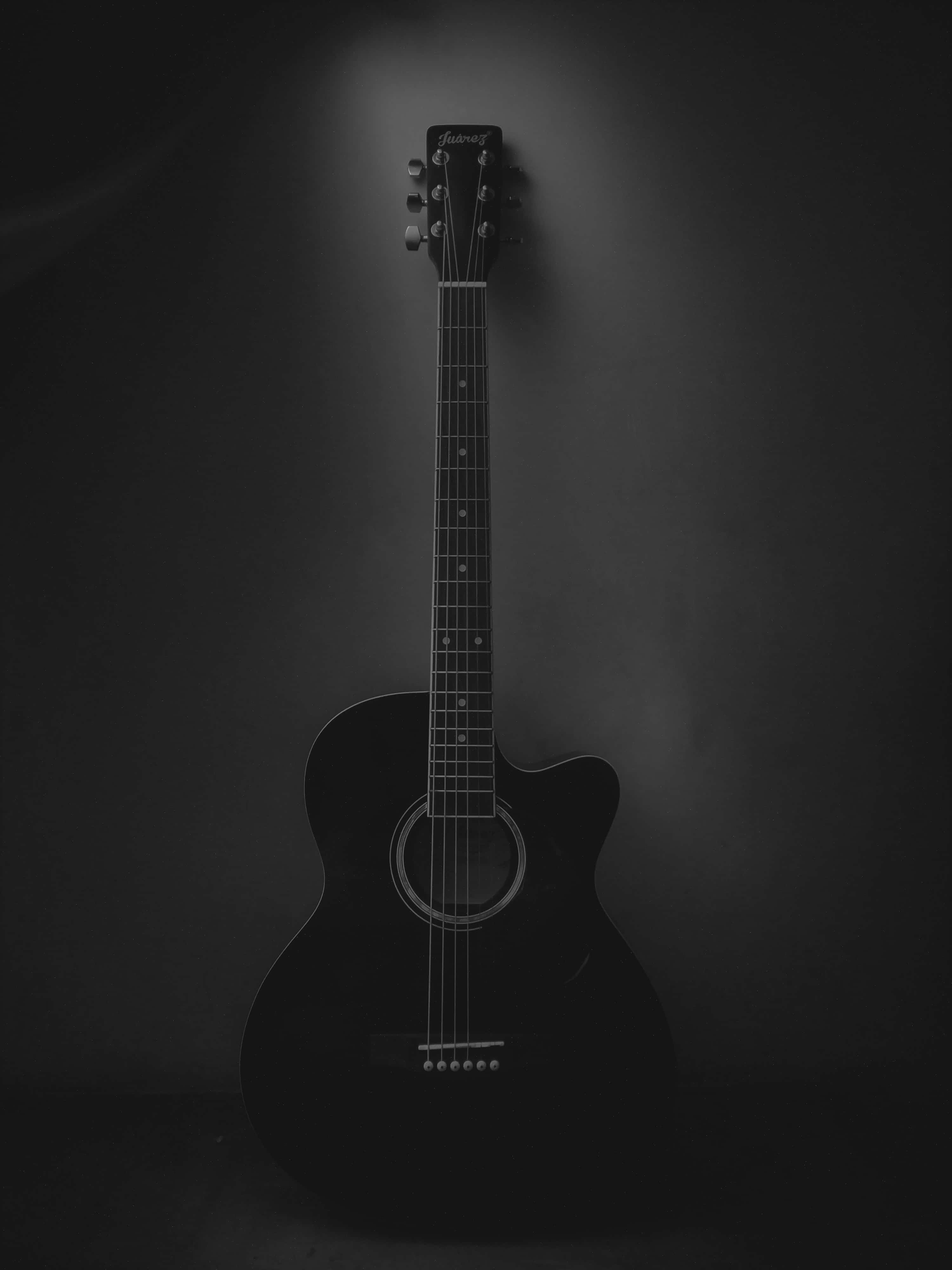 Acoustic Guitar 桌布 壁紙 — 一把黑色木吉他垂直豎立在黑暗背景前，由單一聚光燈照射，形成戲劇性的陰影。