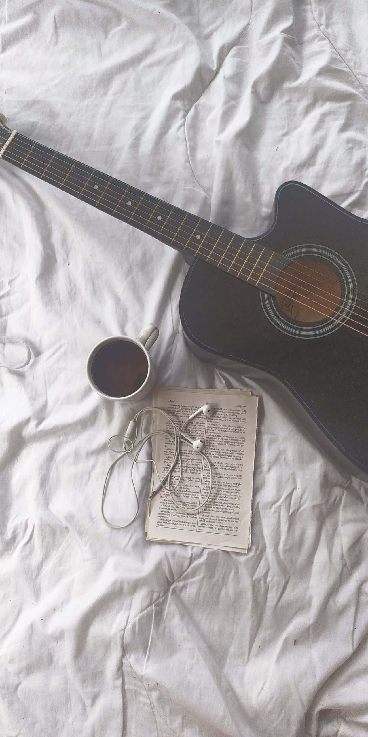 Guitar Aesthetic 桌布 壁紙 — 一把黑色木吉他放在皺巴巴的白色床單上，旁邊有咖啡杯、耳塞和一本打開的書。