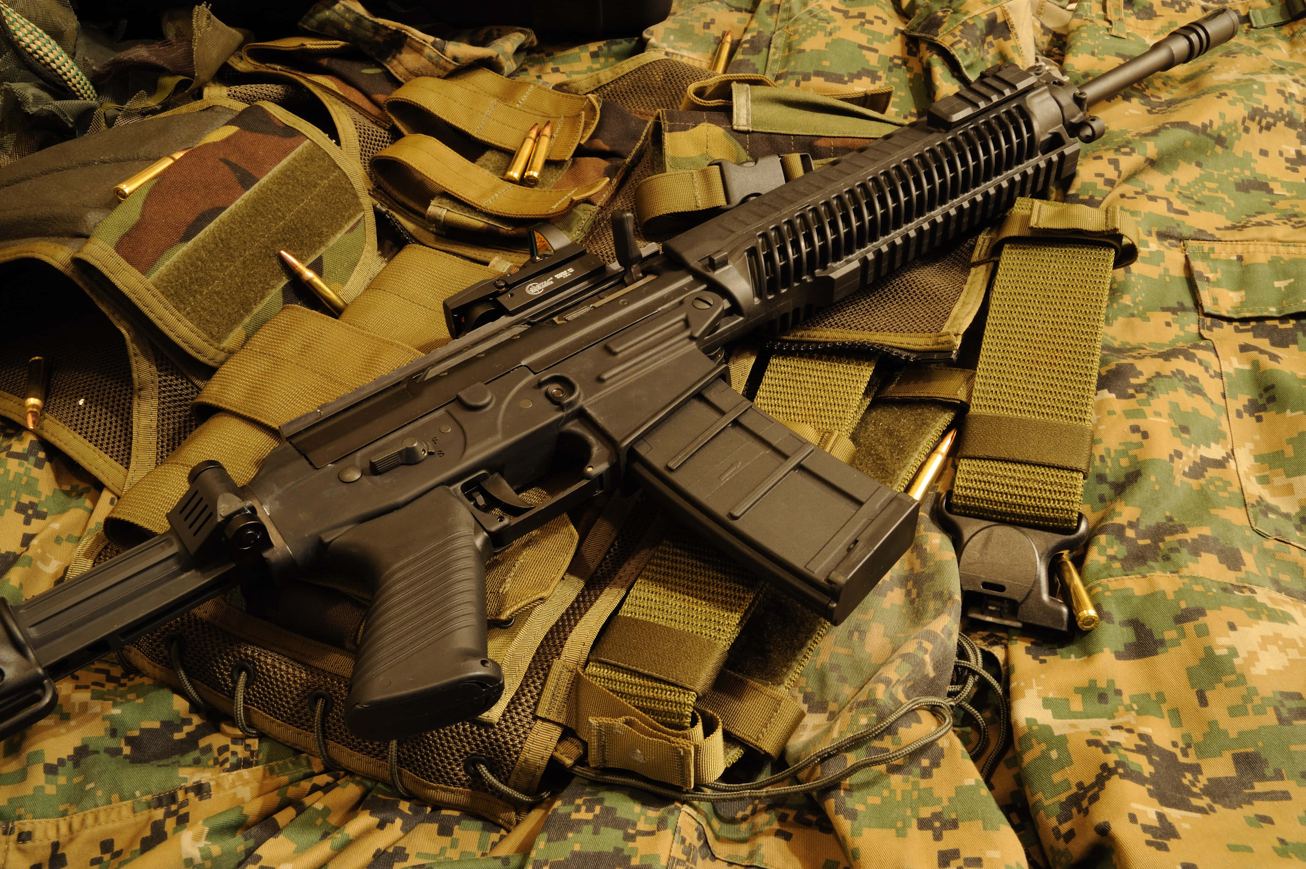 Rifle 桌布 壁紙 — 一把黑色突擊步槍放在迷彩裝備上，周圍散落著子彈和戰術帶，在溫暖燈光下。