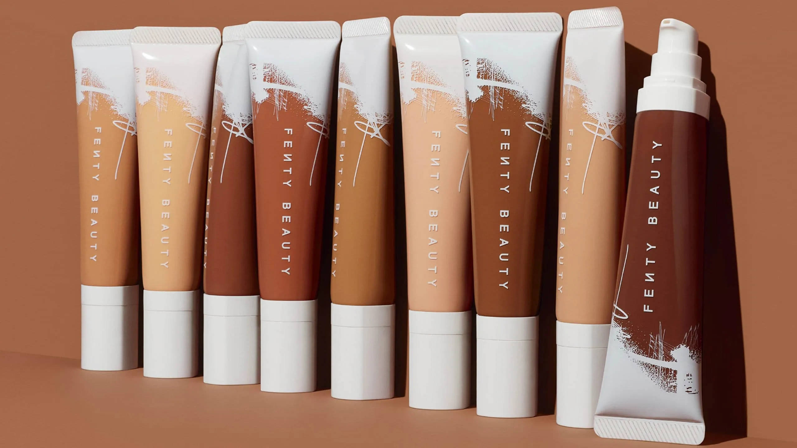 Fenty 桌布 壁紙 — 一排Fenty Beauty粉底管，呈現各種溫暖色調，整齊排列在陶土色背景前。