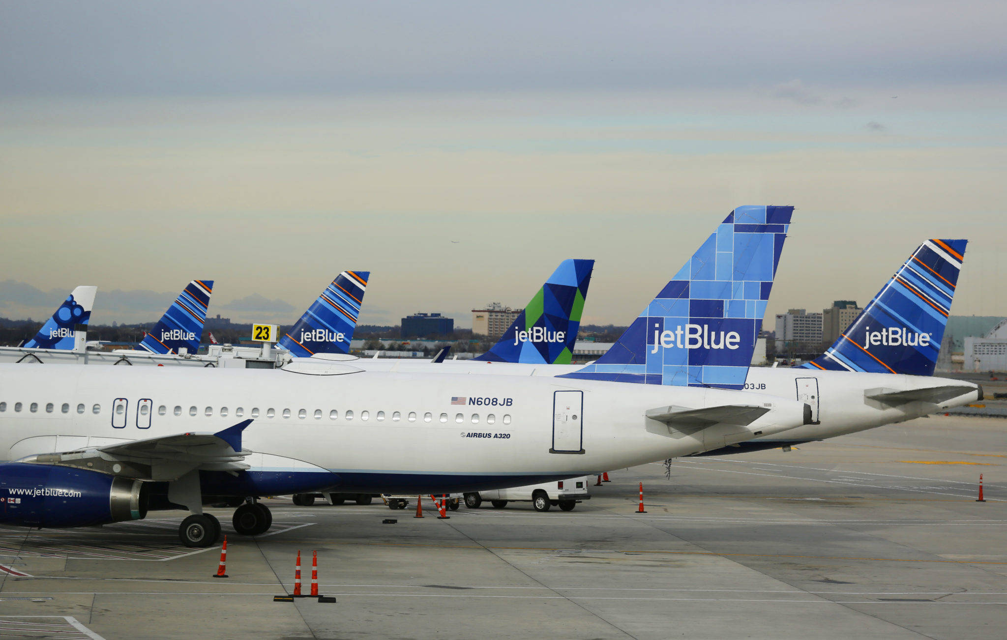 Plane 桌布 壁紙 — 一排JetBlue空中巴士A320飛機停放在機場停機坪上，天空多雲，遠處有城市建築物。