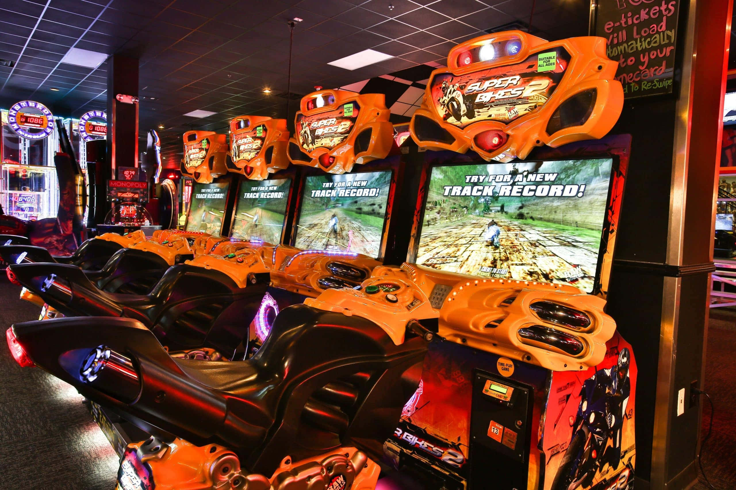 Arcade Games 桌布 壁紙 — 一排亮橙色賽車街機，配備發光螢幕和方向盤，位於燈光昏暗的遊戲廳。
