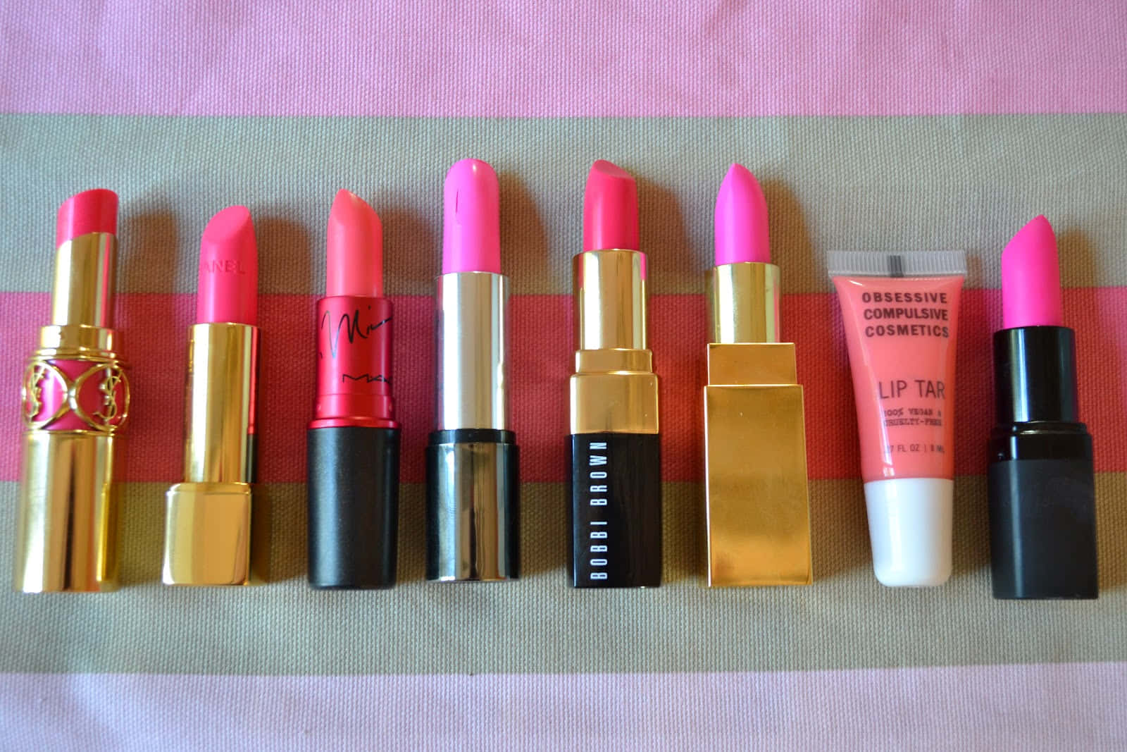 Pink Lipstick 桌布 壁紙 — 一排來自奢華和獨立品牌的鮮豔粉紅色唇膏和唇彩，整齊地排列在條紋布料背景上。