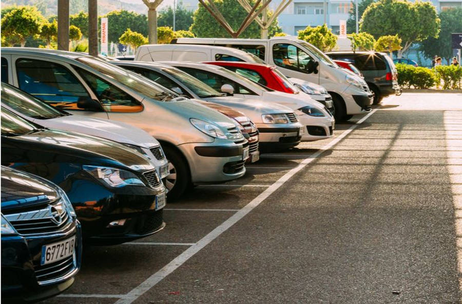 汽車 桌布 壁紙 — 一排停放在陽光普照的停車場的汽車，背景有樹木和建築物。