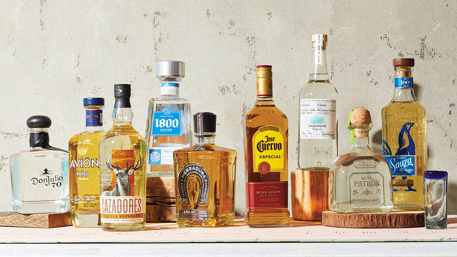 alcohol 桌布 壁紙 — 一排優質龍舌蘭酒瓶，標籤各異，琥珀色液體，展示在木製杯墊上，背景為紋理牆壁。