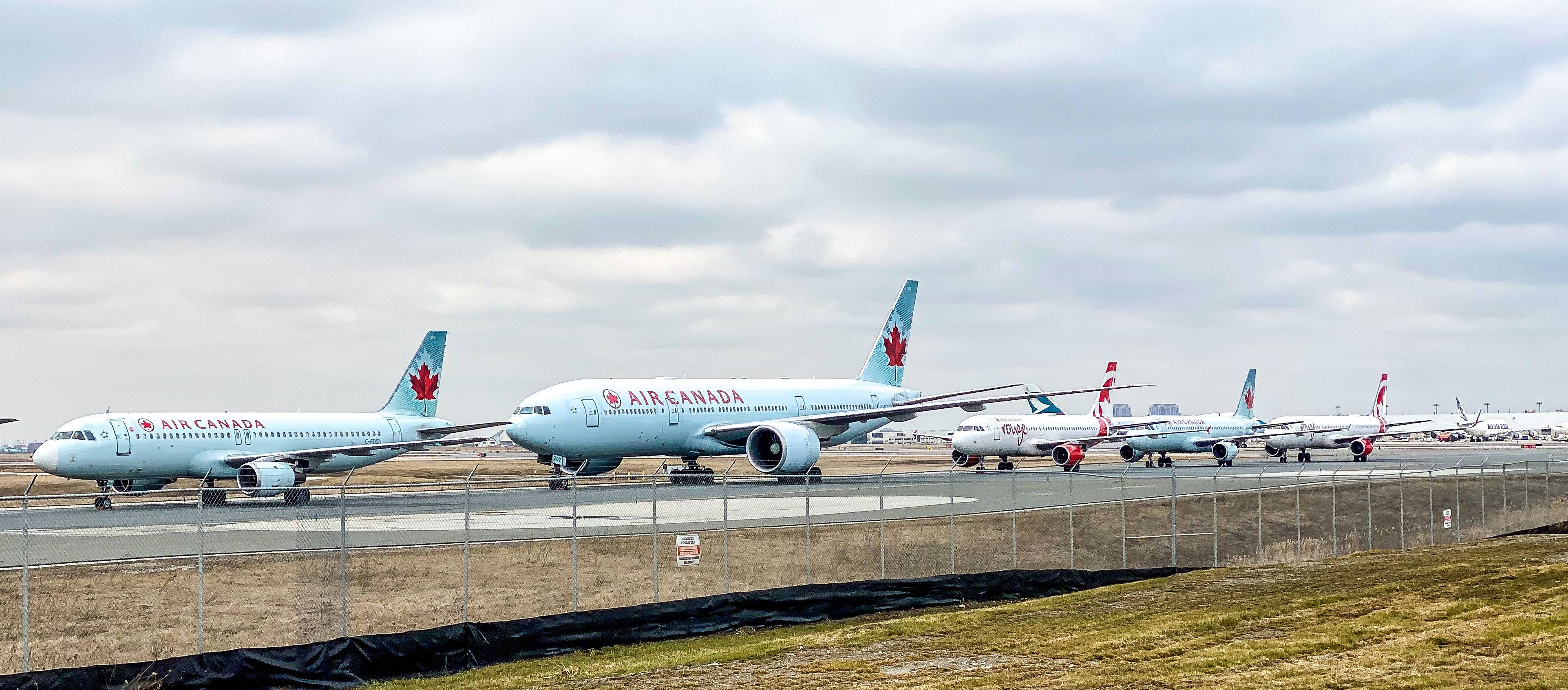 飛機 桌布 壁紙 — 一排加拿大航空飛機停放在機場停機坪上，天空多雲，前景有鐵絲網圍欄。