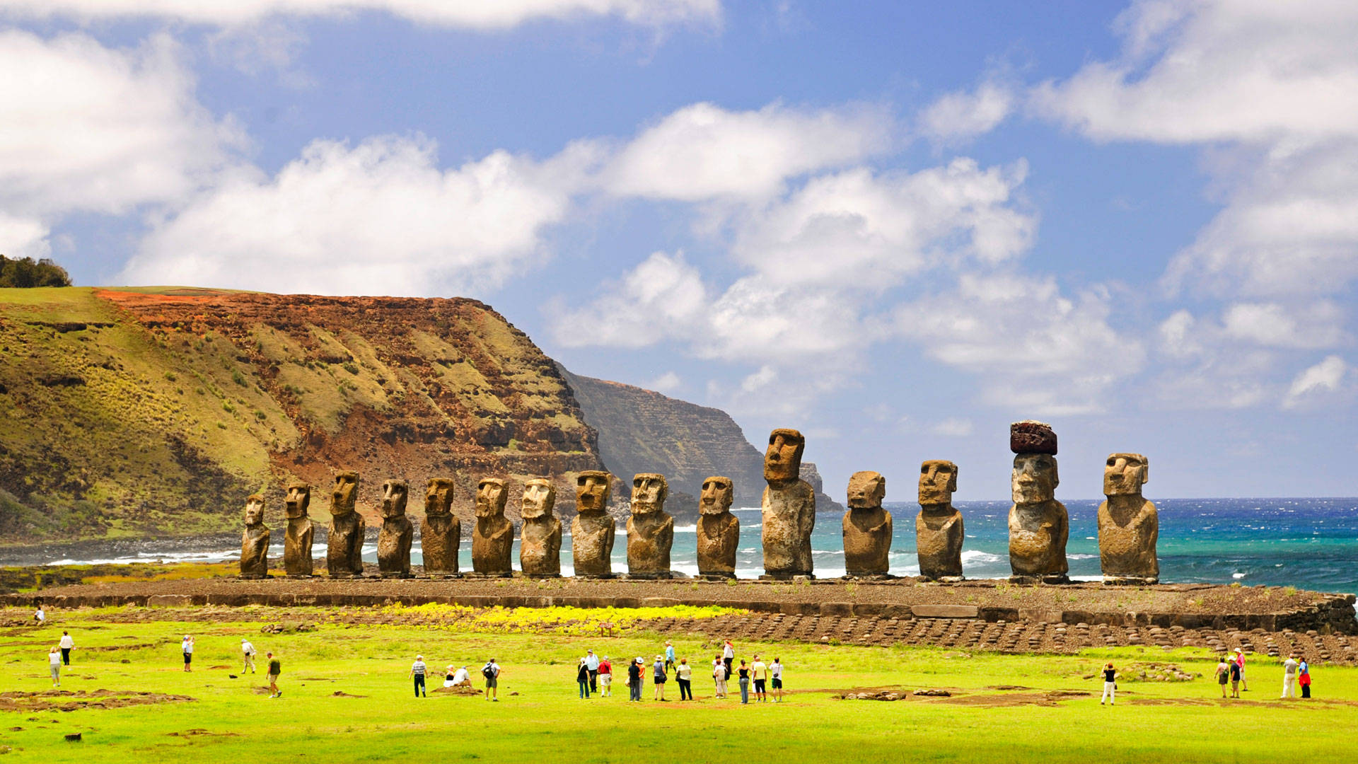 South America 桌布 壁紙 — 一排古老的摩艾石像莊嚴地矗立在長滿青草的懸崖上，俯瞰著碧綠色的海洋，天空湛藍，白雲朵朵。