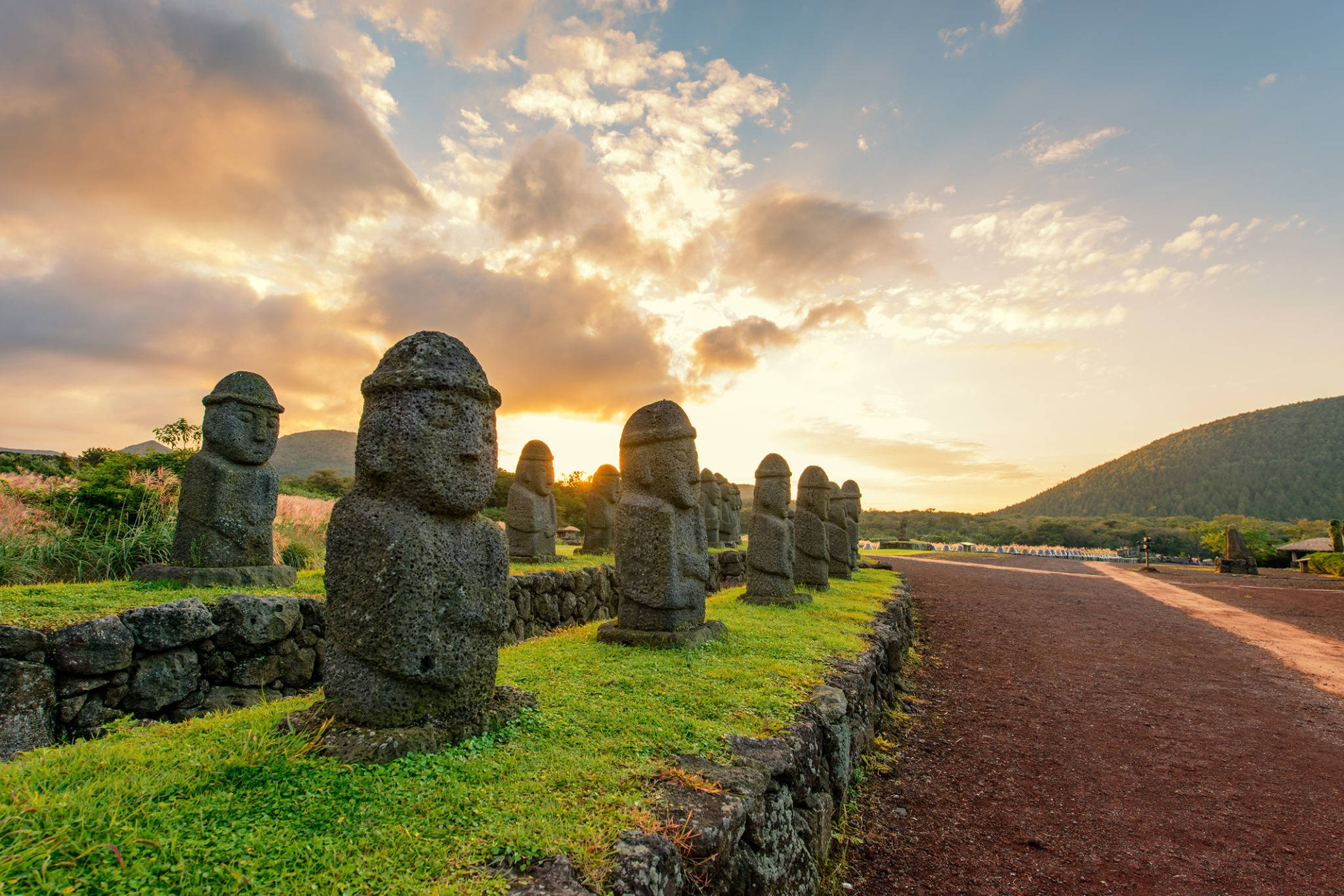 Jeju 桌布 壁紙 — 一排古老的石像摩艾雕像莊嚴地矗立在日落時分的草地上，溫暖的金色光芒照亮了她們風化的臉龐。