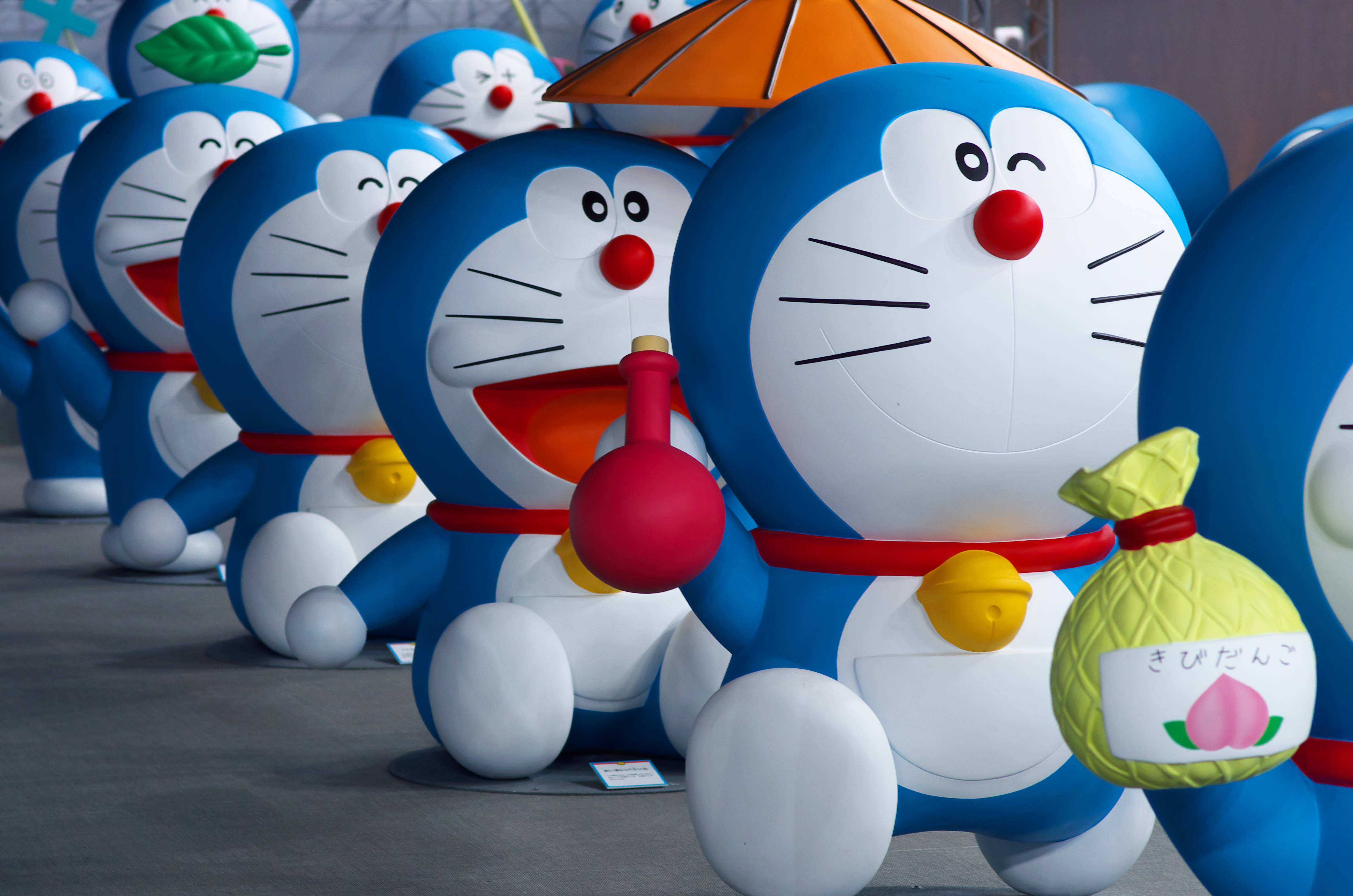 Doraemon 桌布 壁紙 — 一排大型藍色哆啦A夢雕像手持紅色氣球和桃子形狀的袋子，在公共展覽中愉快地微笑。