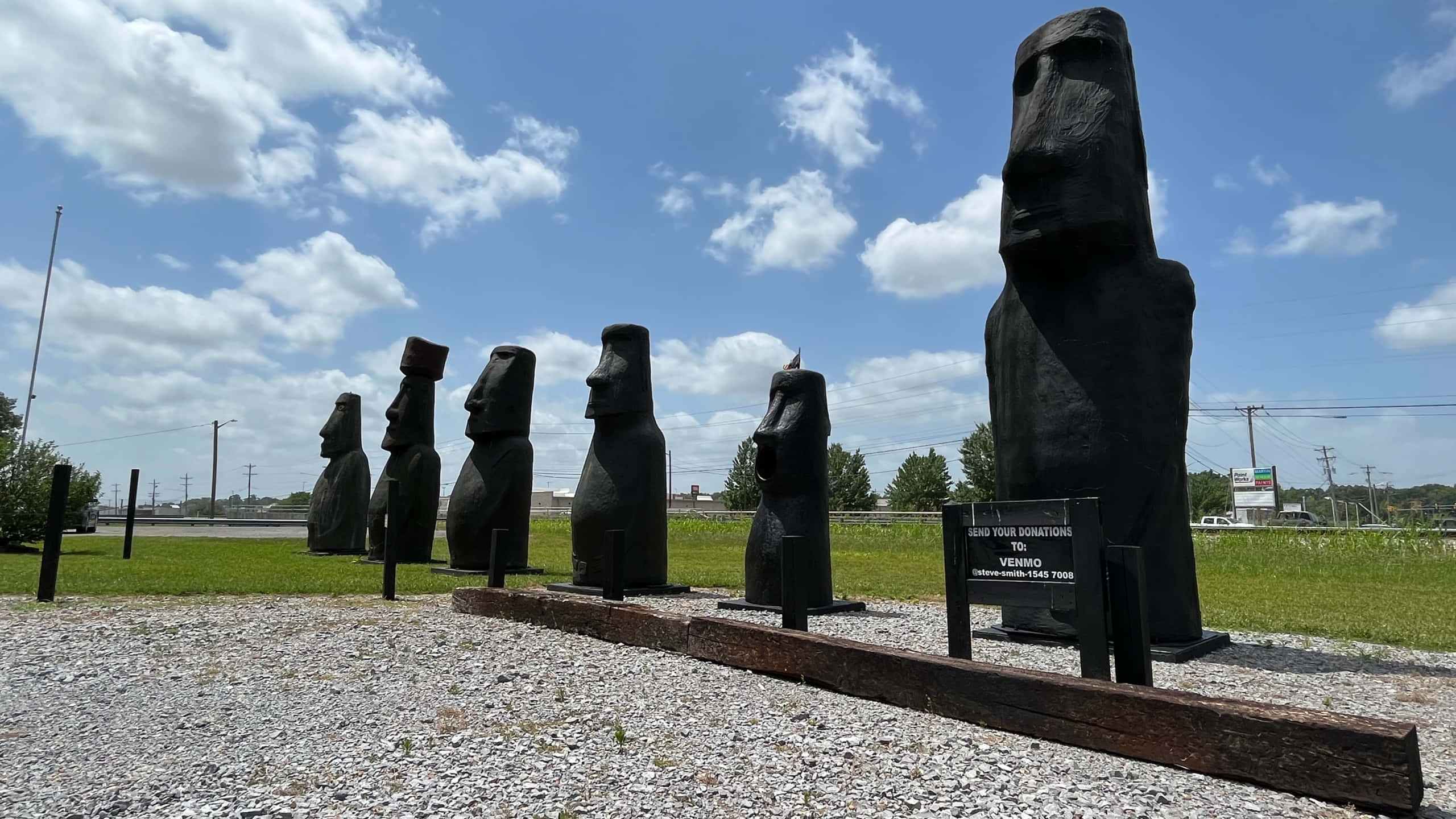 statue 桌布 壁紙 — 一排大型黑色摩艾石像矗立在礫石上，上方是湛藍的天空，飄著蓬鬆的白雲。