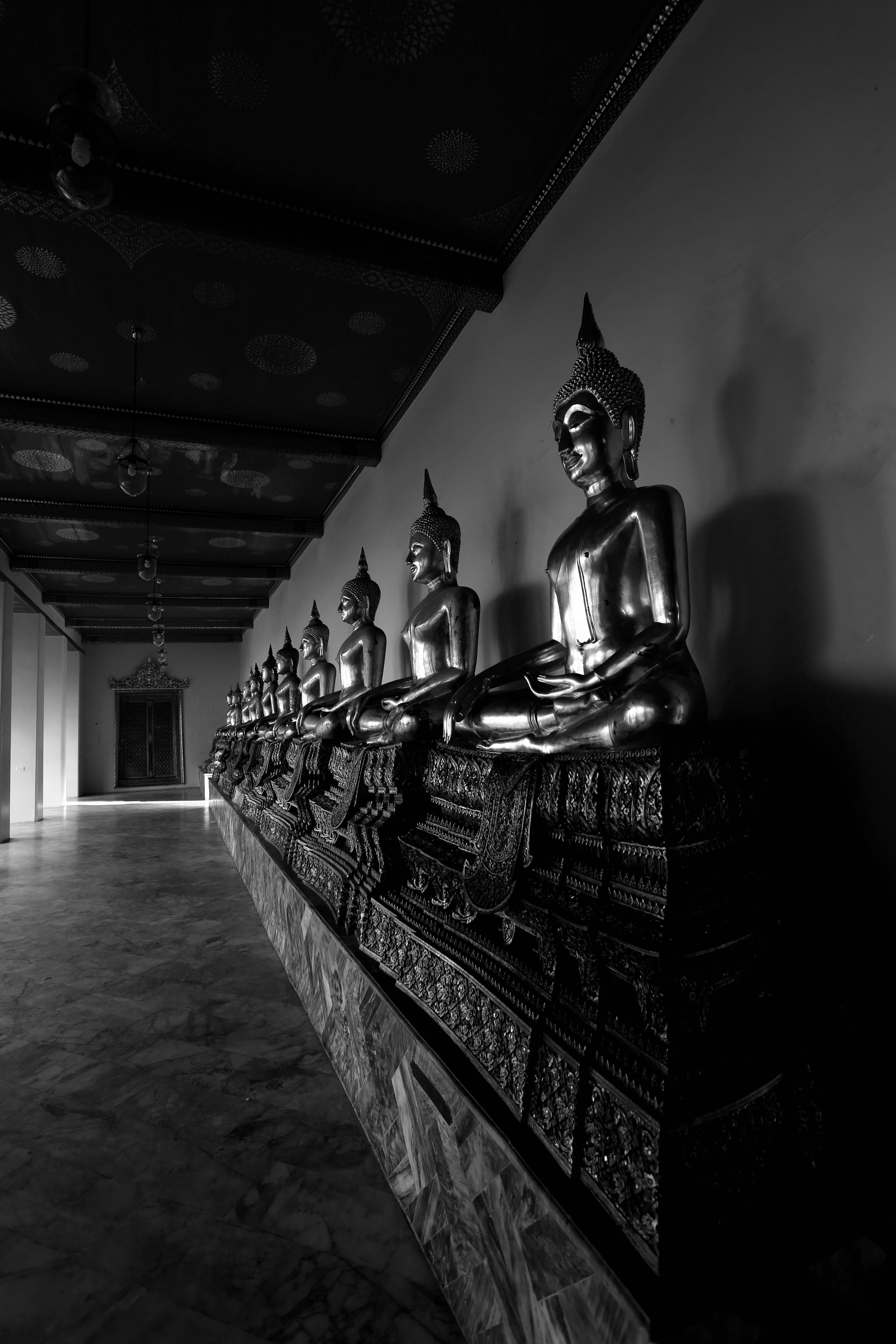 black buddha 桌布 壁紙 — 一排寧靜的佛像以單色呈現，沿著寺廟走廊排列，天花板圖案華麗。