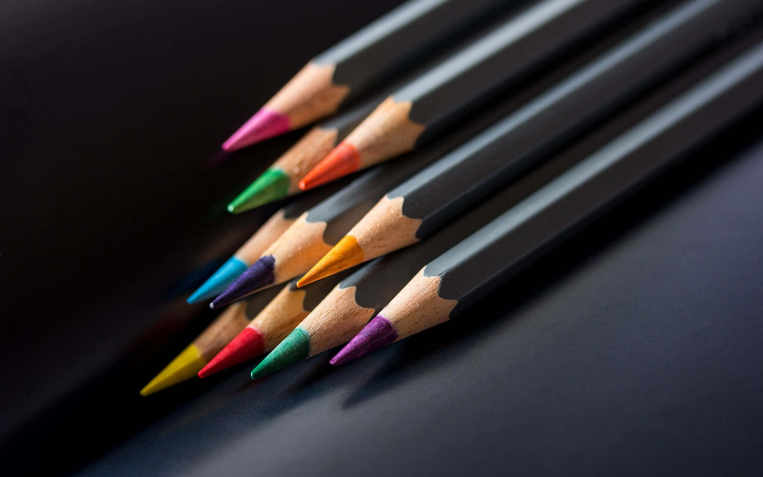 pencil 桌布 壁紙 — 一排對角線的削尖彩色鉛筆，帶有鮮豔的筆尖，在柔和光線下放置在深色表面上。