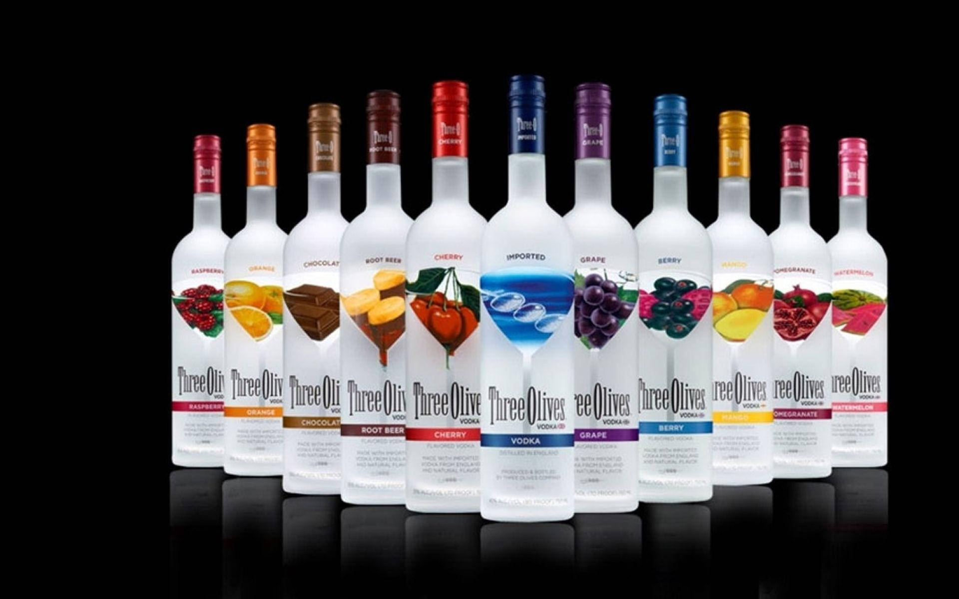 alcohol 桌布 壁紙 — 一排帶有水果和口味標籤的 Twelve Three Olives 伏特加酒瓶整齊地排列在帶有反射表面的黑色背景前。