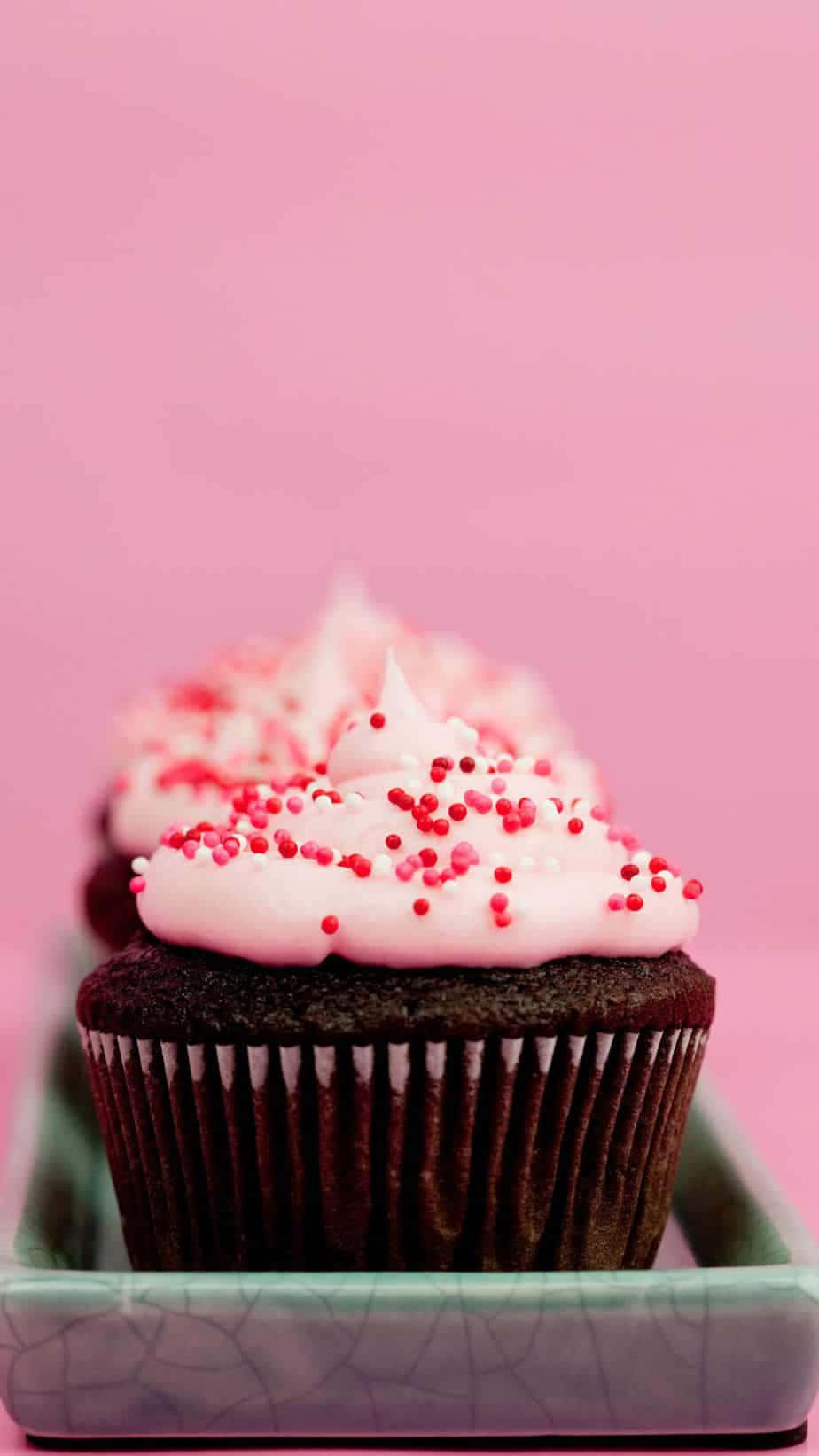 Cute Cupcake 桌布 壁紙 — 一排帶有粉紅色糖霜和紅色糖粒的巧克力杯子蛋糕，放在一個有裂紋的綠色盤子上，背景是柔和的粉紅色。