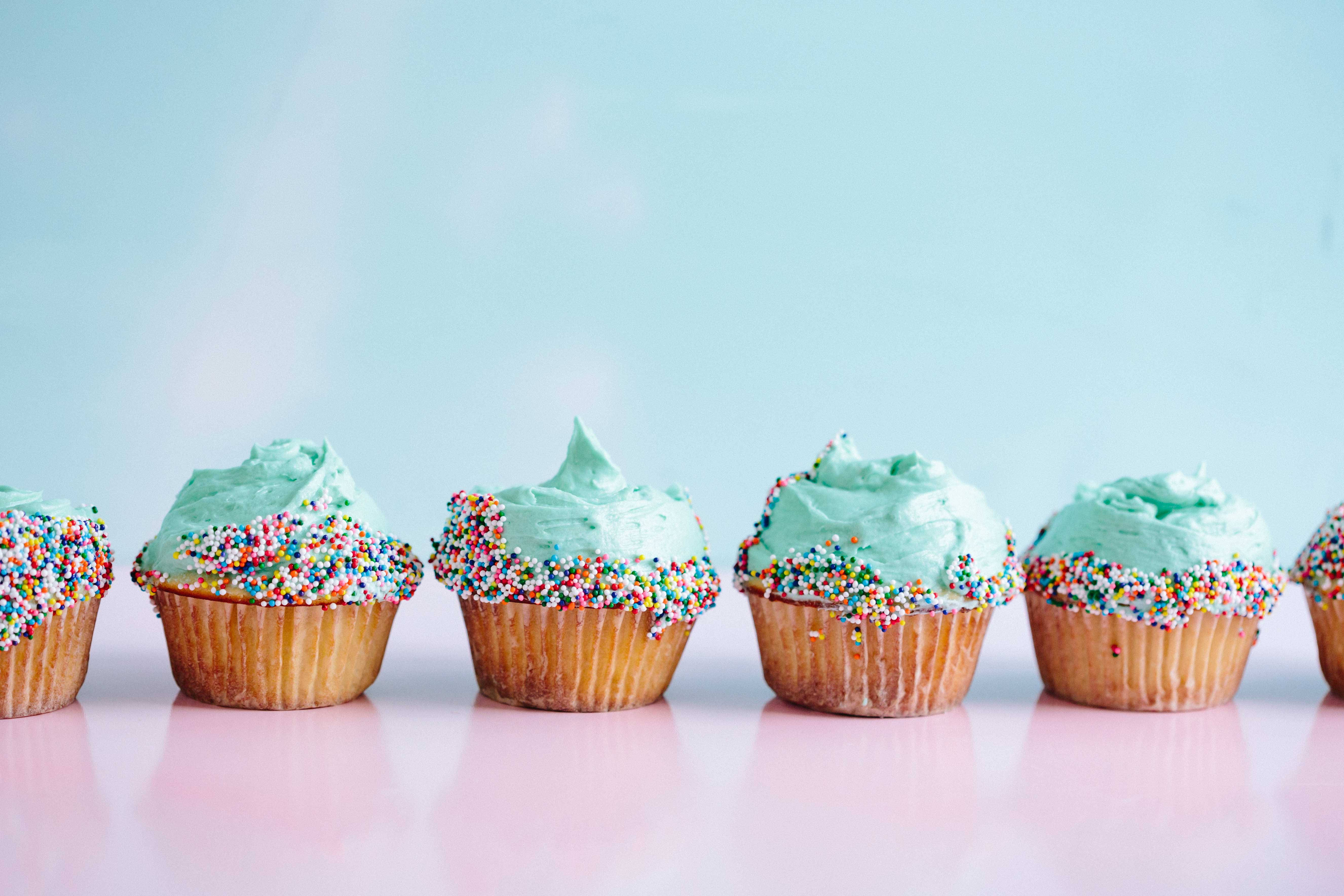 Cupcake 桌布 壁紙 — 一排帶有薄荷綠色糖霜和彩色糖粒的杯子蛋糕，放在粉紅色表面上，背景是柔和的藍色。