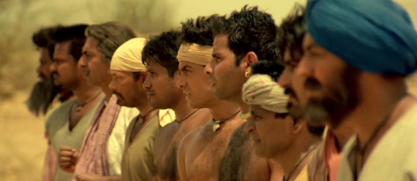 Aamir Khan 桌布 壁紙 — 一排強壯的男人戴著頭巾和頭飾，在溫暖金色光線下的塵土飛揚沙漠景觀中莊嚴地站著。