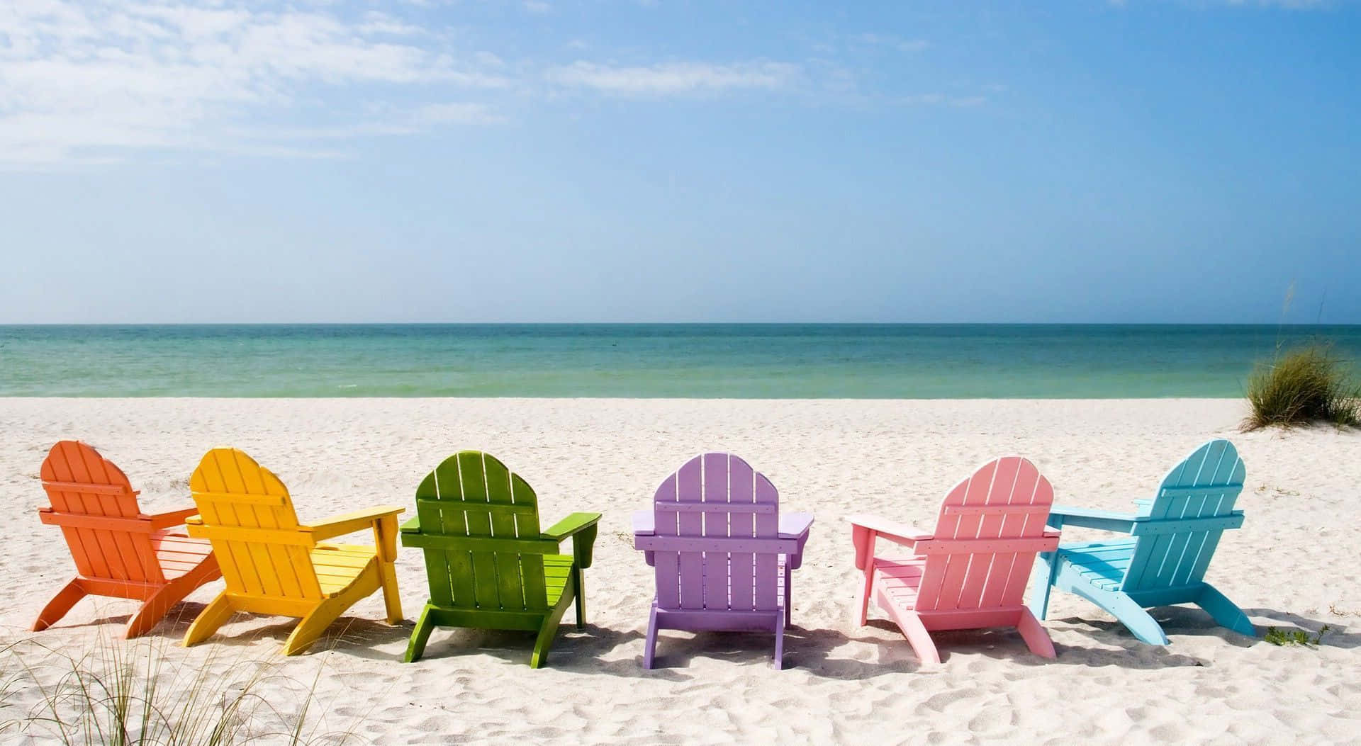 HD Beach 桌布 壁紙 — 一排彩虹色的 Adirondack 椅子面向藍綠色的海洋，在明亮的白色沙灘上，天空清澈湛藍。