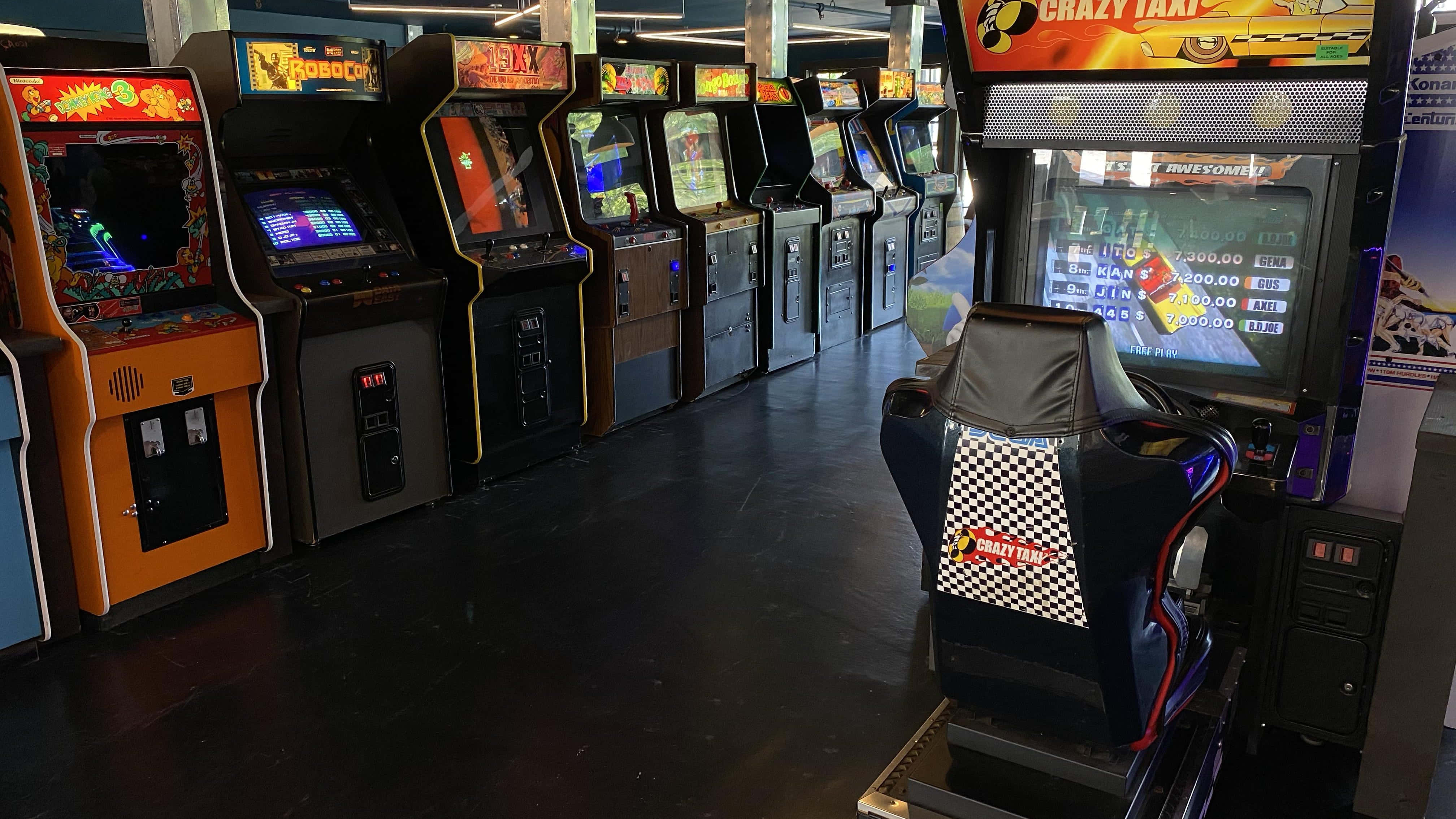 Arcade Games 桌布 壁紙 — 一排復古街機，帶有發光的螢幕和色彩繽紛的機櫃，排列在燈光昏暗的遊戲廳中。