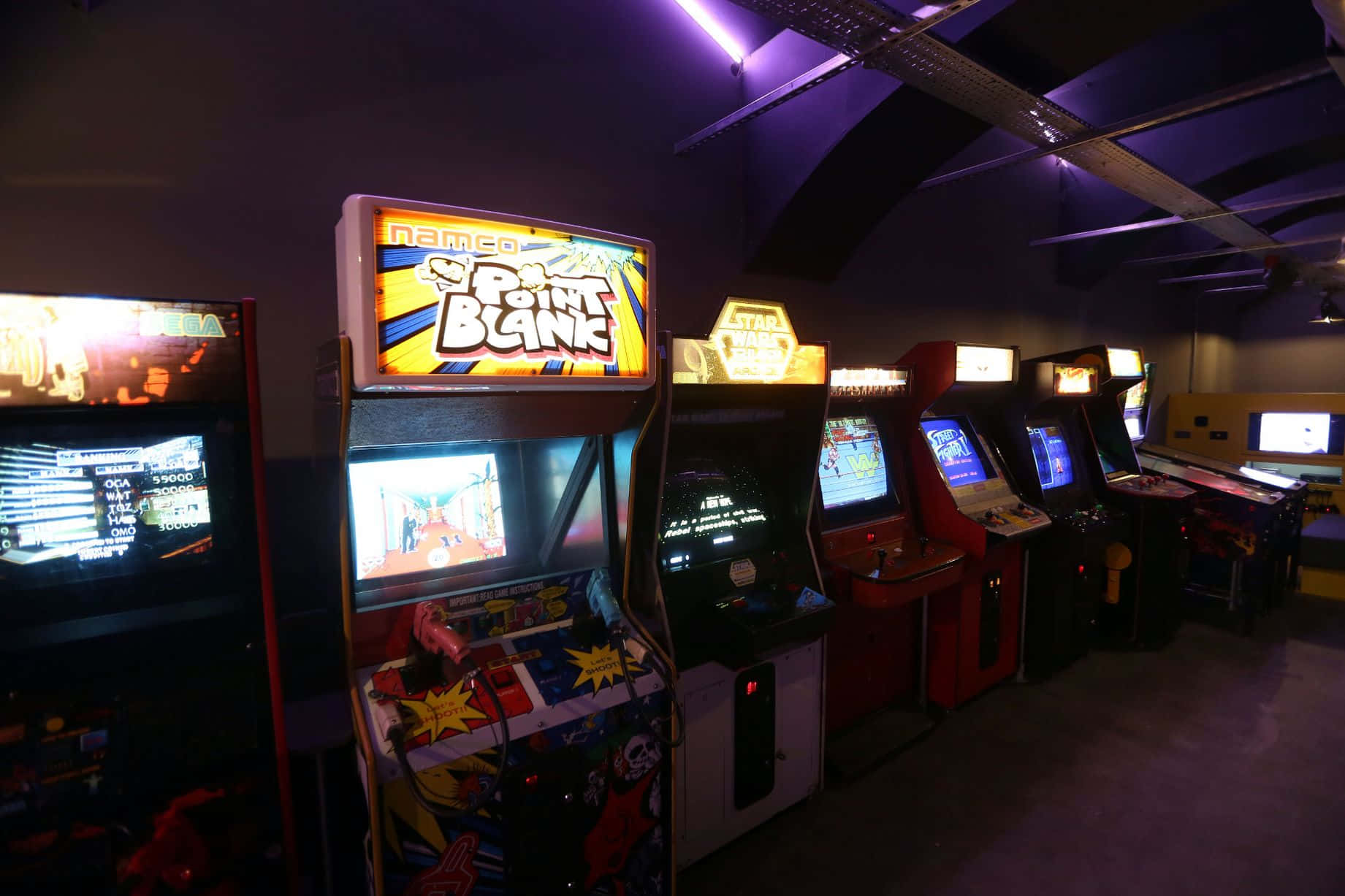 Arcade Games 桌布 壁紙 — 一排復古街機在昏暗的復古遊戲廳中，在紫色燈光下閃耀。