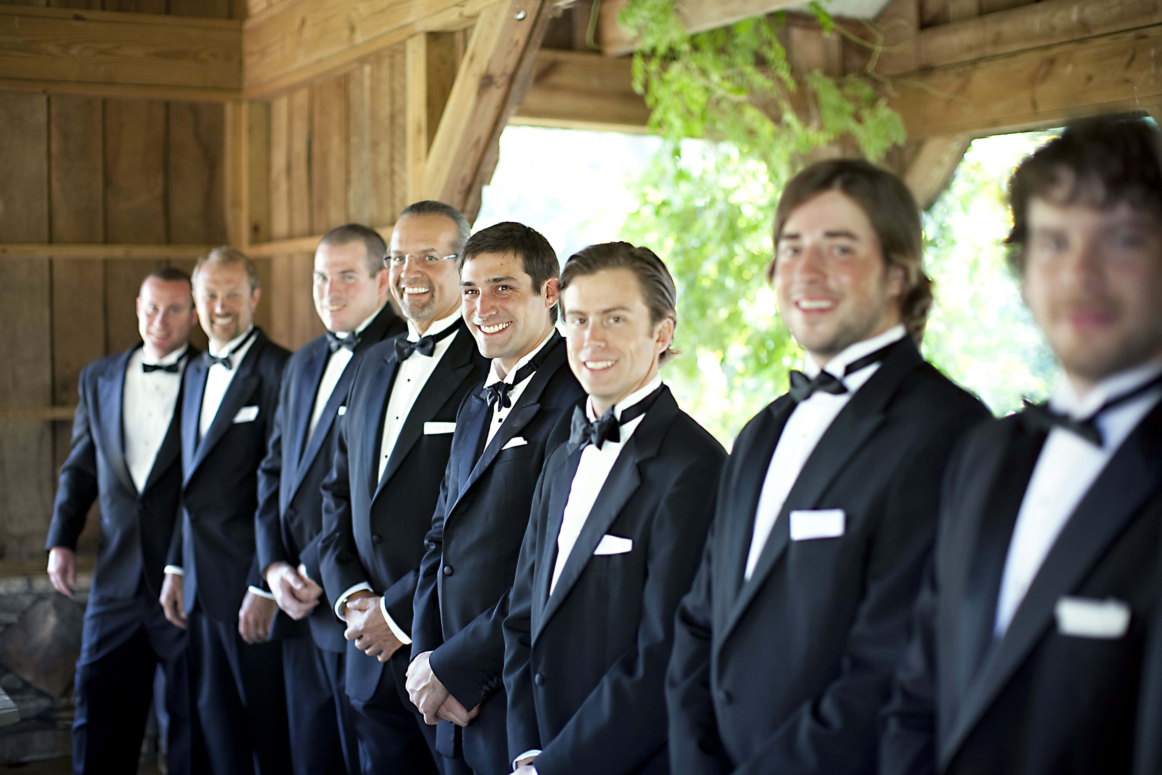 Groomsmen 桌布 壁紙 — 一排微笑的伴郎穿著黑色燕尾服，站在一個質樸的木製涼亭下，身後有綠色樹葉。