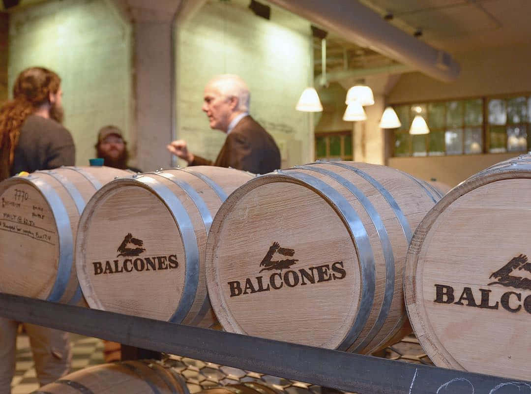 政治 桌布 壁紙 — 一排排 Balcones 威士忌木桶放在燈光溫暖的釀酒廠內，背景中人們在交談。