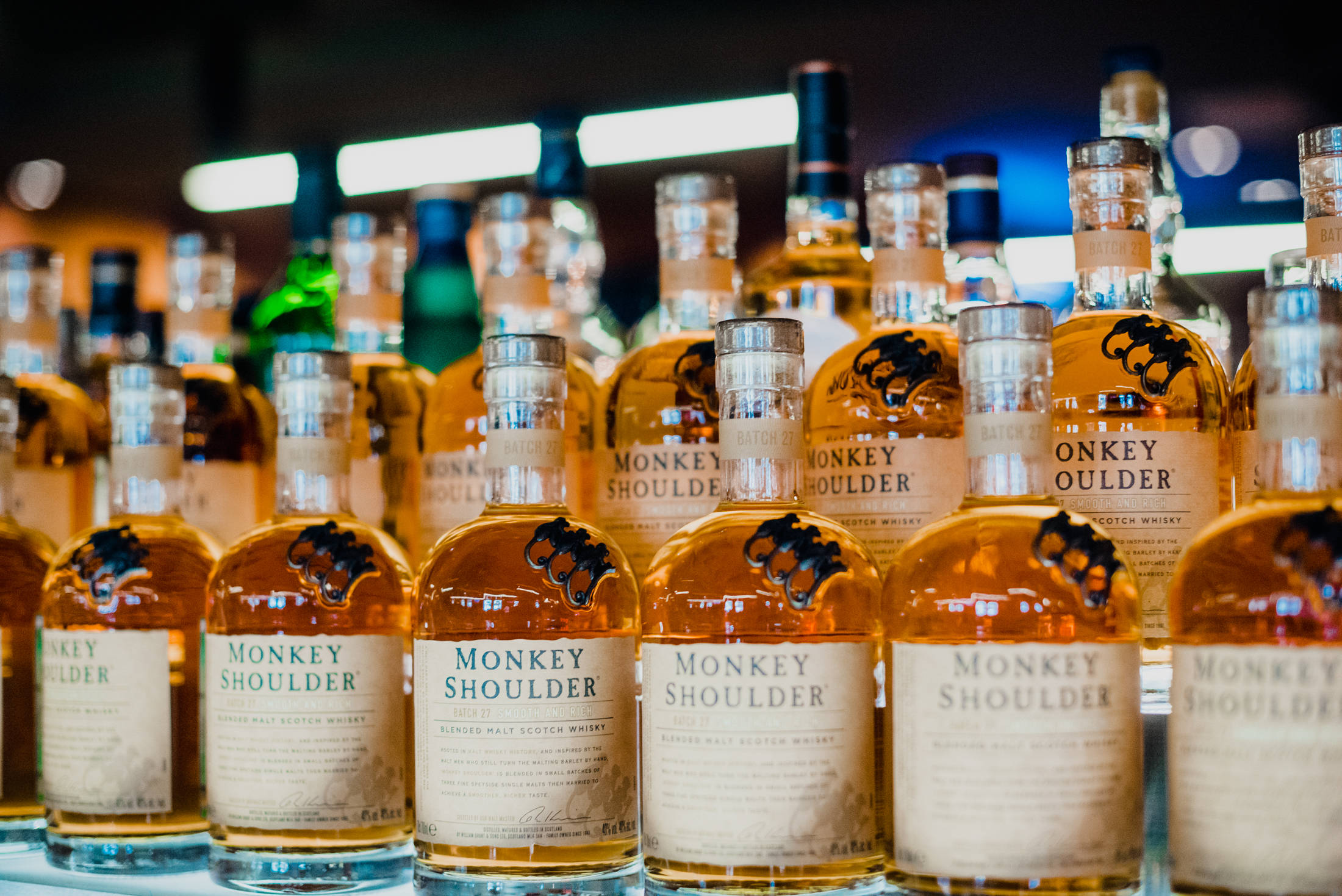 alcohol 桌布 壁紙 — 一排排 Monkey Shoulder 威士忌酒瓶在時尚酒吧環境中，於明亮燈光下閃閃發光。