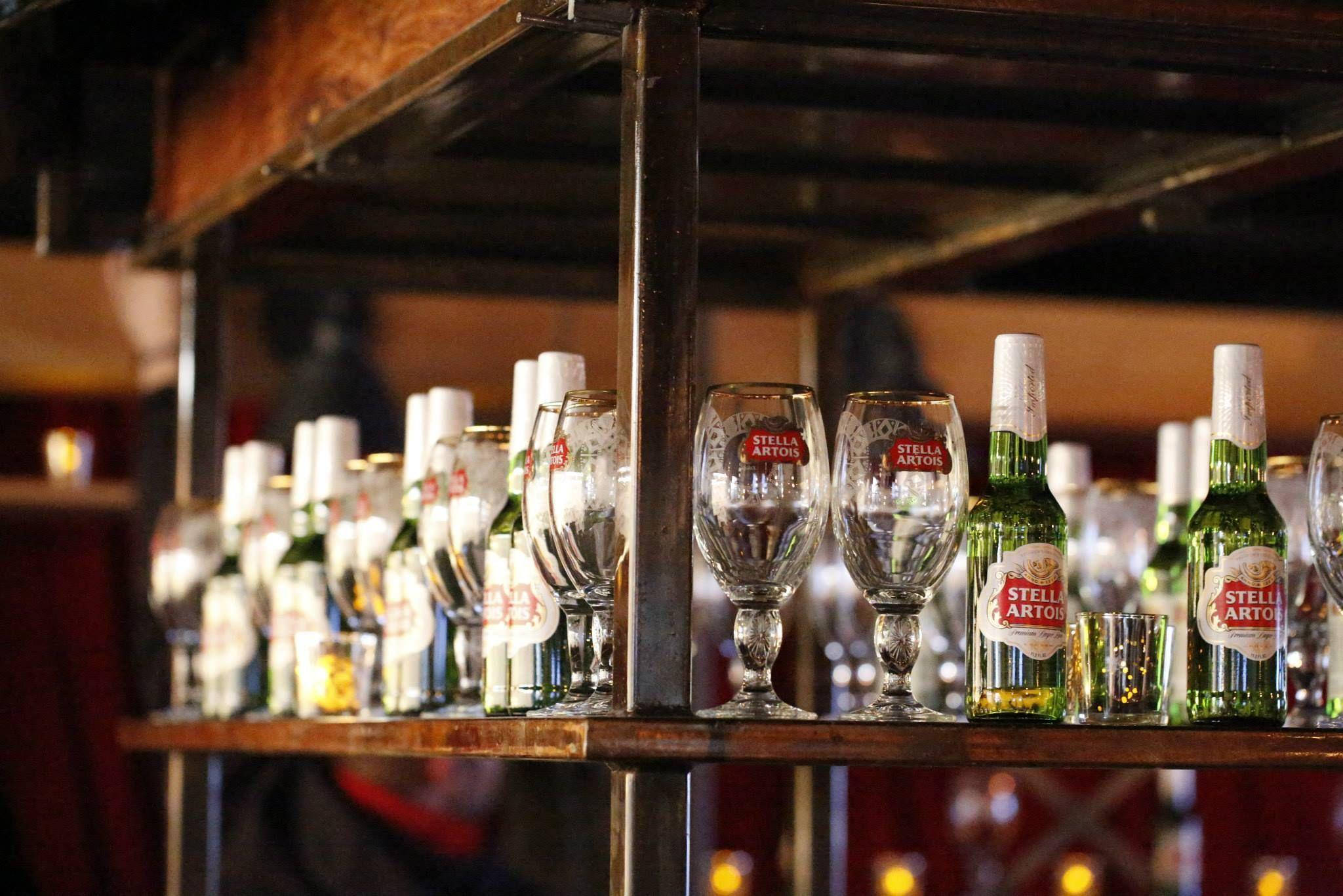 飲料 桌布 壁紙 — 一排排 Stella Artois 啤酒瓶和酒杯在木架上溫暖的酒吧燈光下閃閃發光。