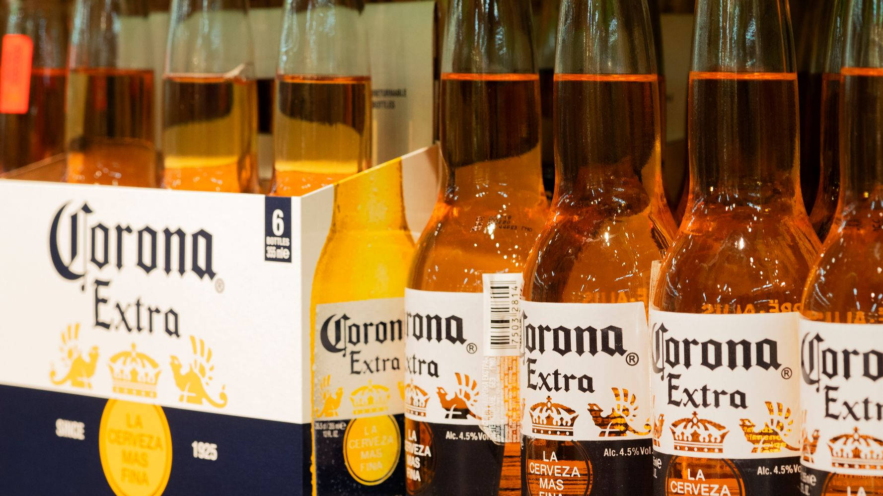 corona 桌布 壁紙 — 一排排Corona Extra啤酒瓶，裝有金色液體和白色標籤，旁邊放著一個有品牌的紙箱，在商店環境中。