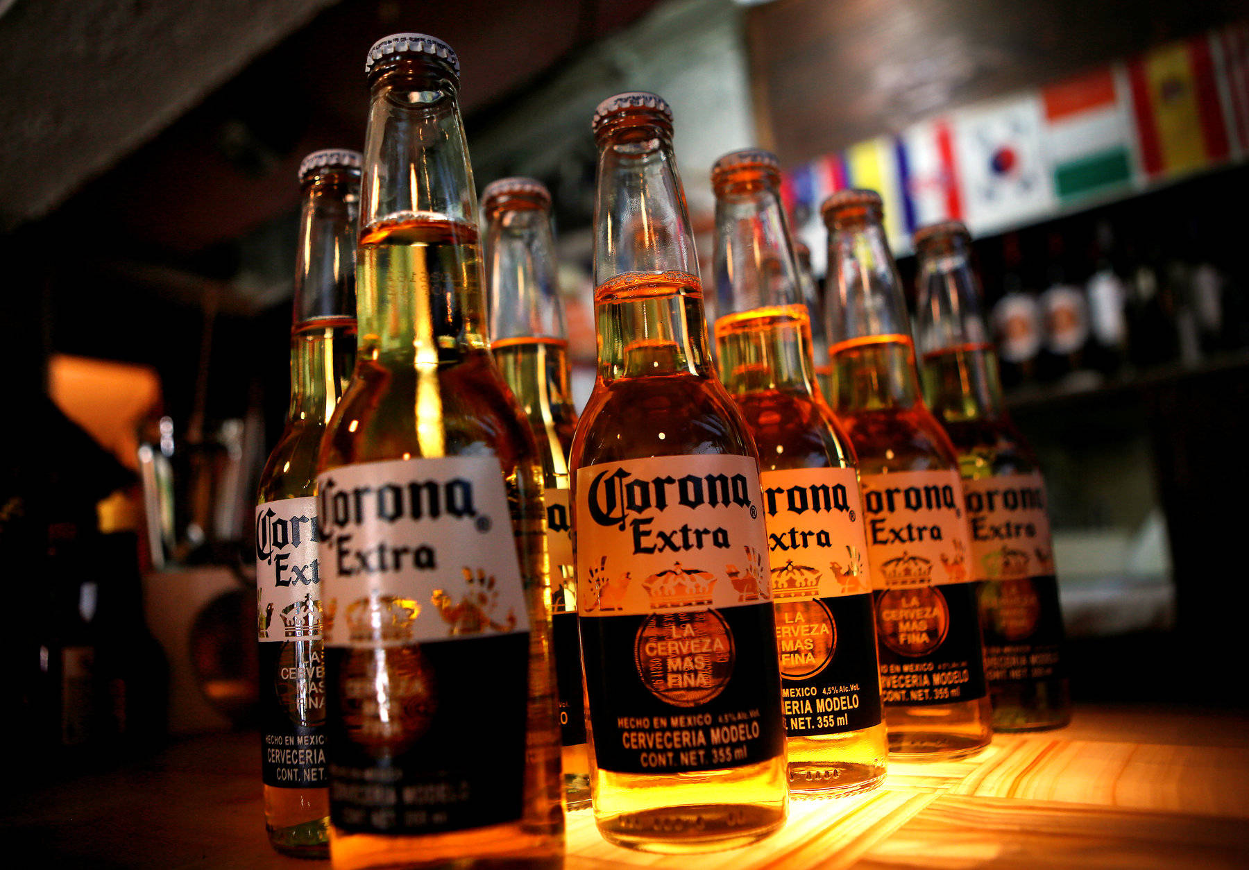 corona 桌布 壁紙 — 一排排Corona Extra啤酒瓶在溫暖的酒吧燈光下閃閃發光，背景可見旗幟。