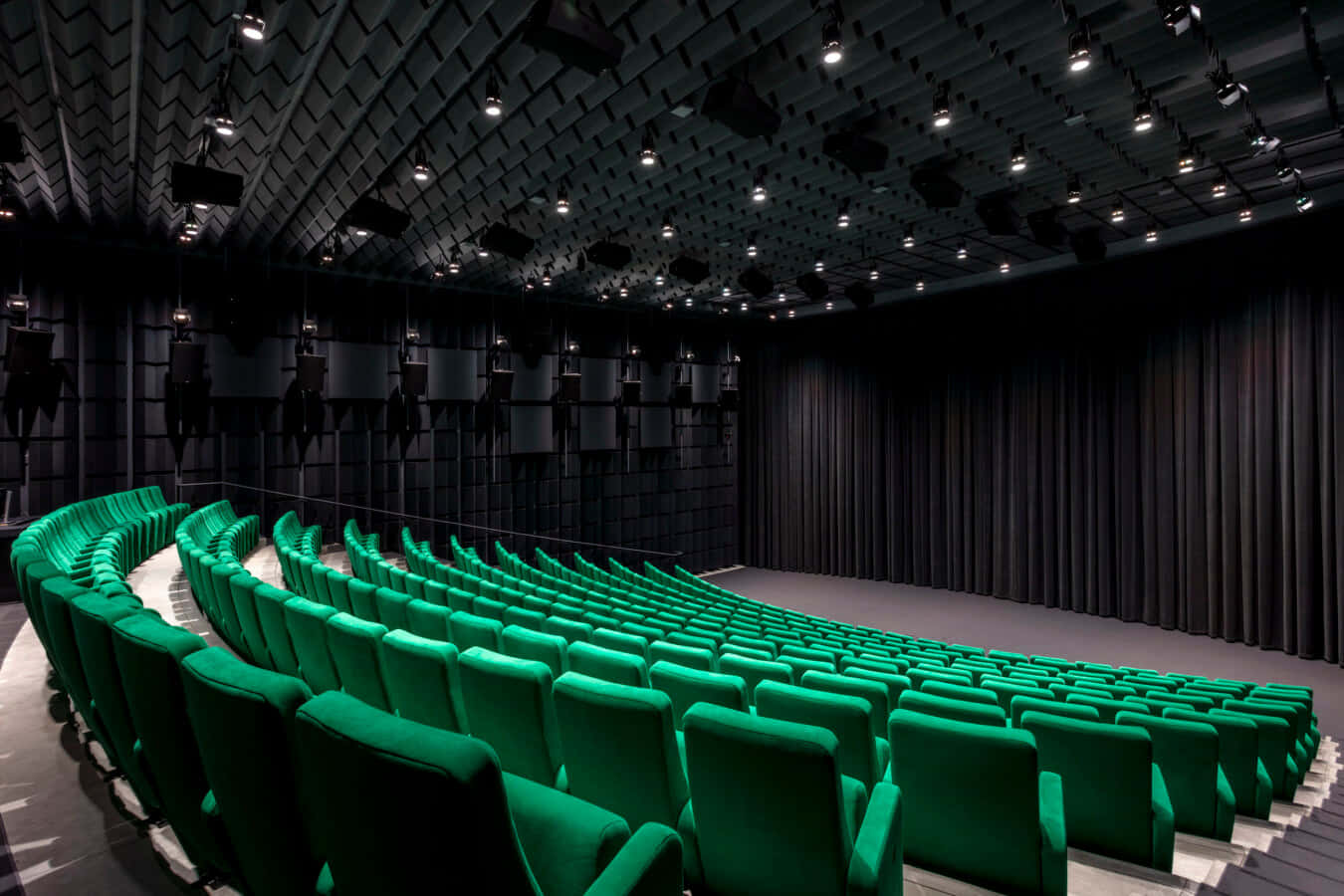 Theater 桌布 壁紙 — 一排排充滿活力的綠色天鵝絨座椅在現代劇院中彎曲向黑暗的舞台，天花板上裝有幾何燈光。