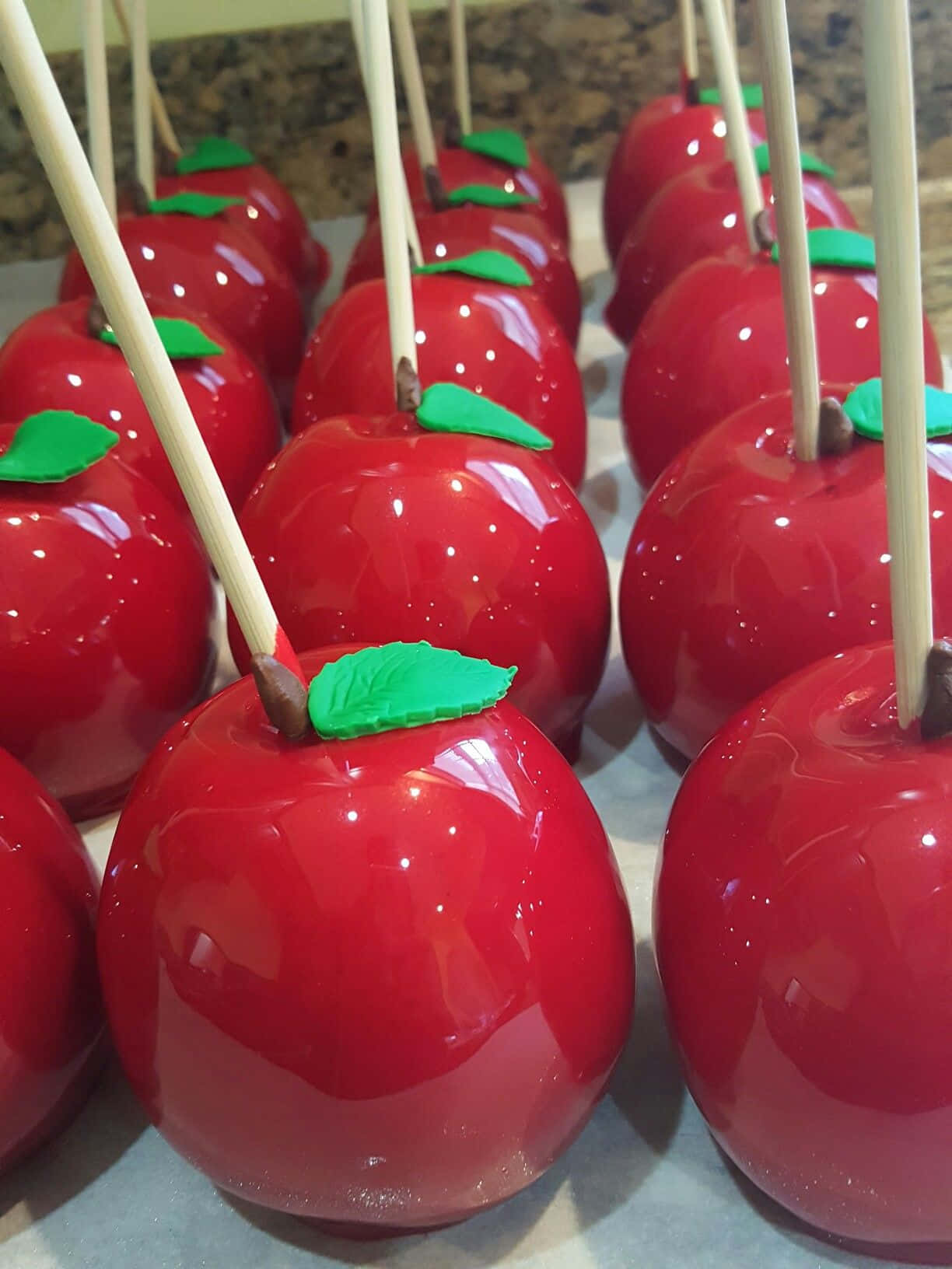Candy Apples 桌布 壁紙 — 一排排光澤的紅色焦糖蘋果，帶有綠葉和棍子，整齊地排列在檯面上。