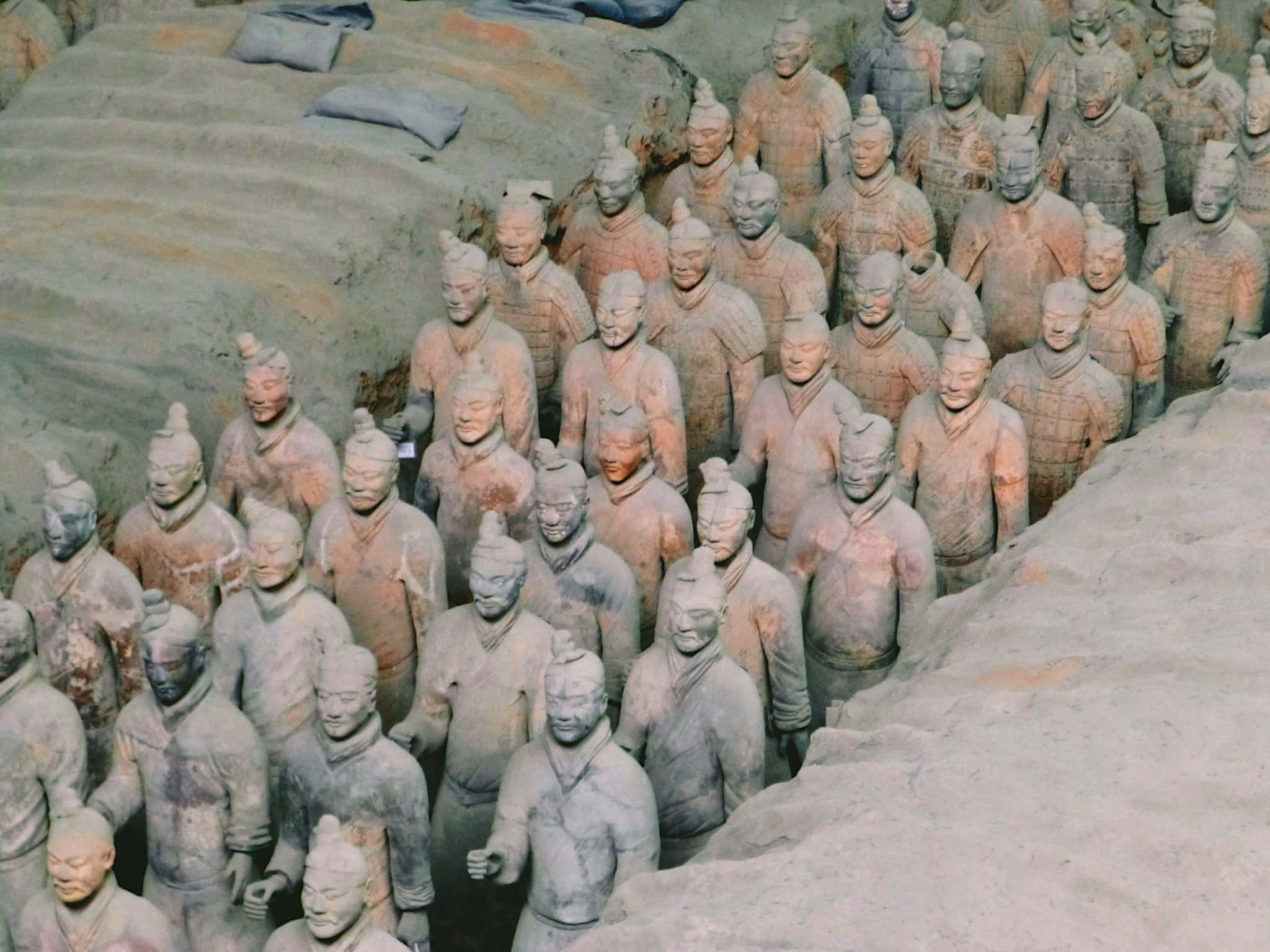 Museum 桌布 壁紙 — 一排排古老的兵馬俑以大地色調矗立在它們的發掘坑中，臉部細節帶有莊嚴的表情。