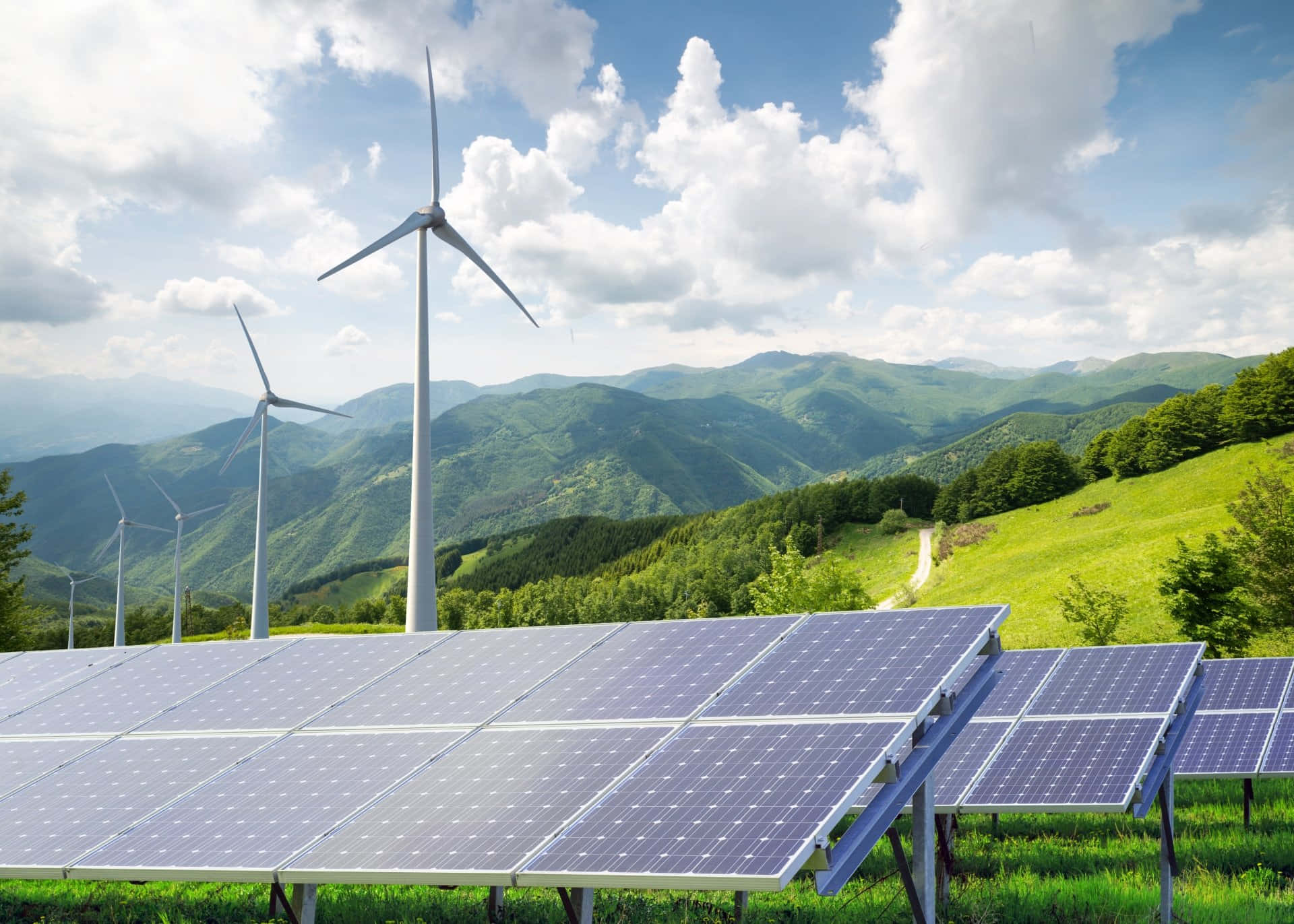 電力 桌布 壁紙 — 一排排太陽能板和風力渦輪機矗立在長滿青草的山坡上，俯瞰著連綿起伏的綠色山脈，天空湛藍，白雲朵朵。